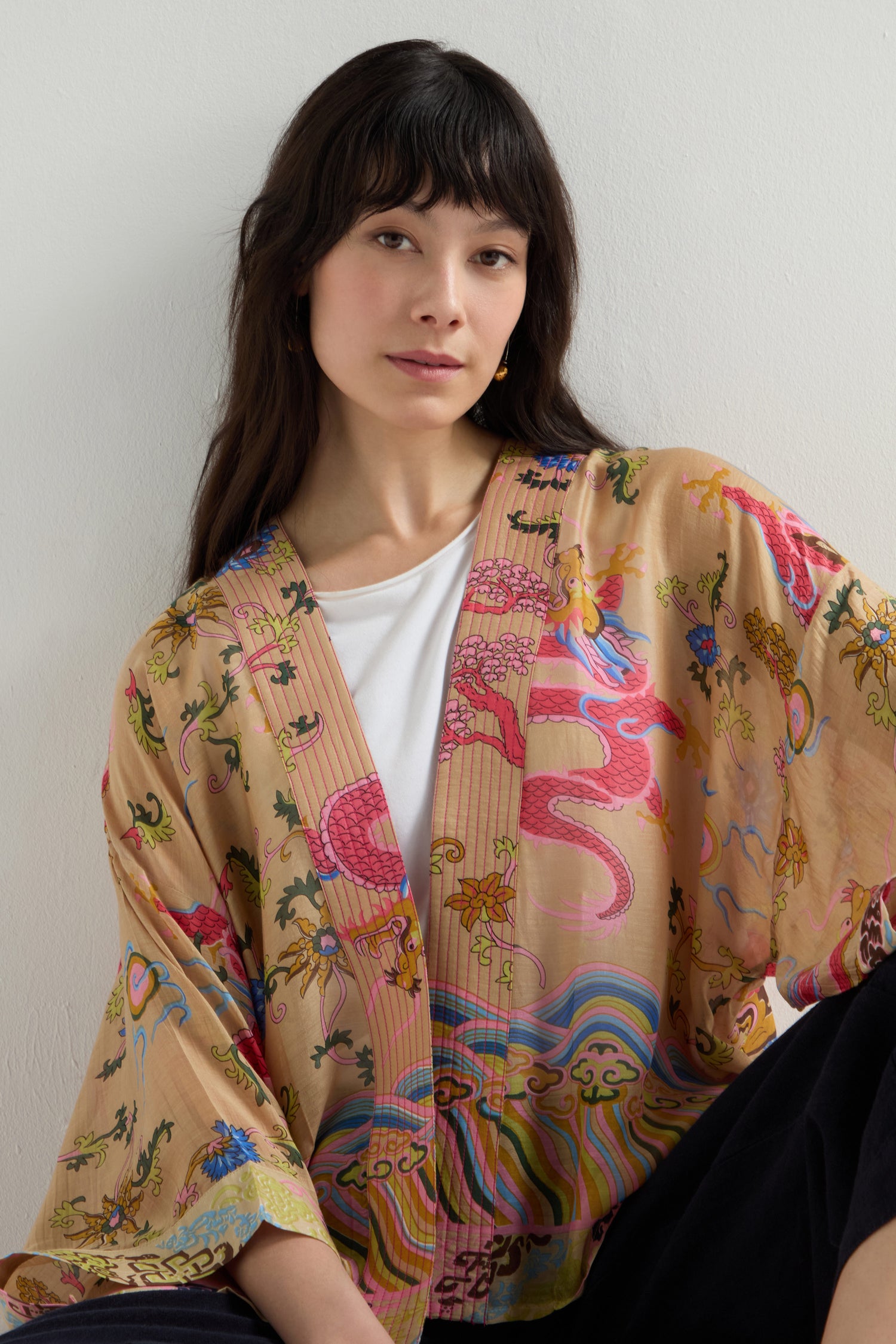 A woman with long dark hair sits against a plain wall, wearing the Dragon Print Kimono over a white top.