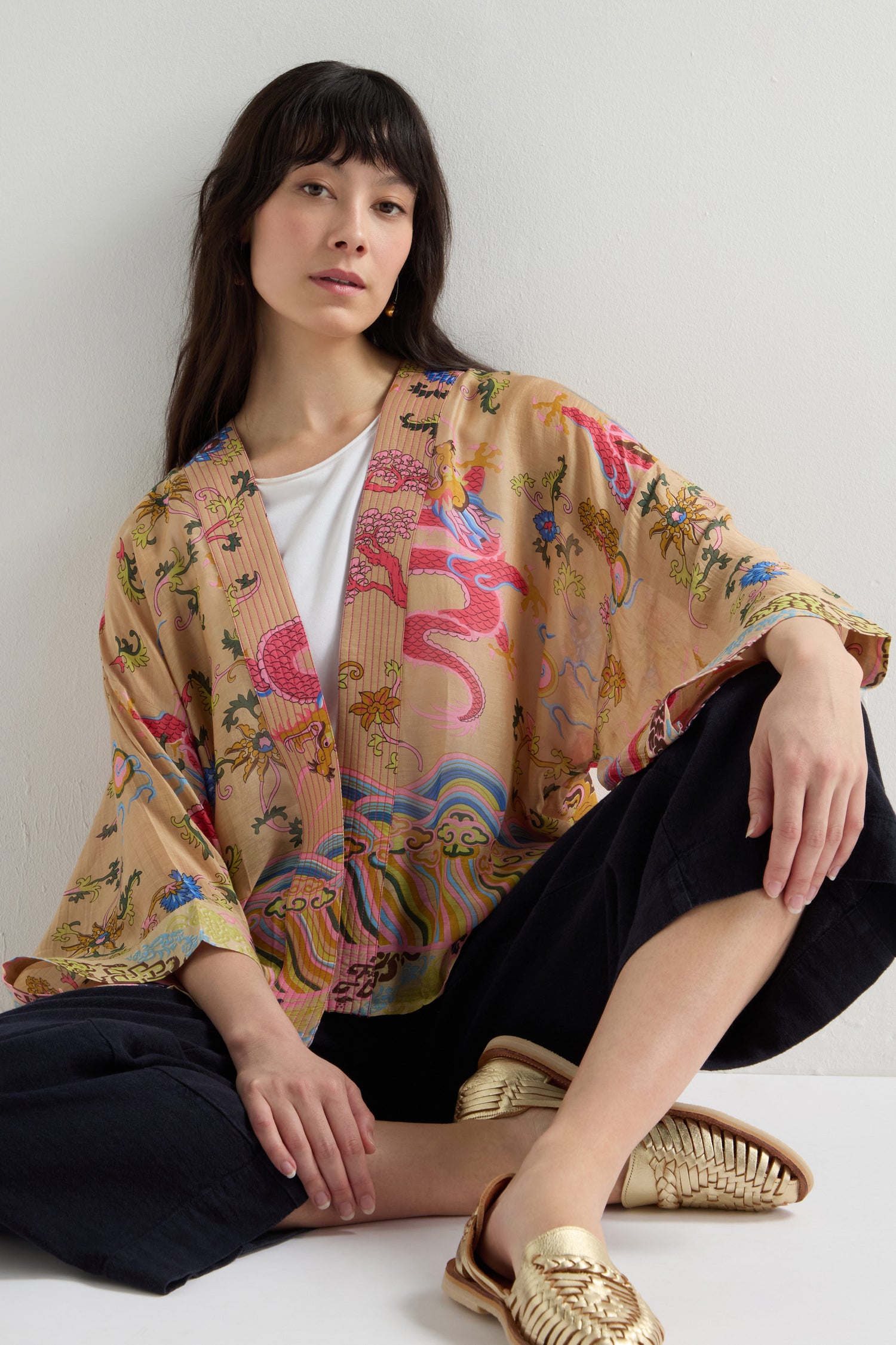 A person sits against a white wall, donning a Dragon Print Kimono with a colorful floral pattern paired with black pants and gold woven shoes. Their pose is relaxed, with one arm resting on a knee.