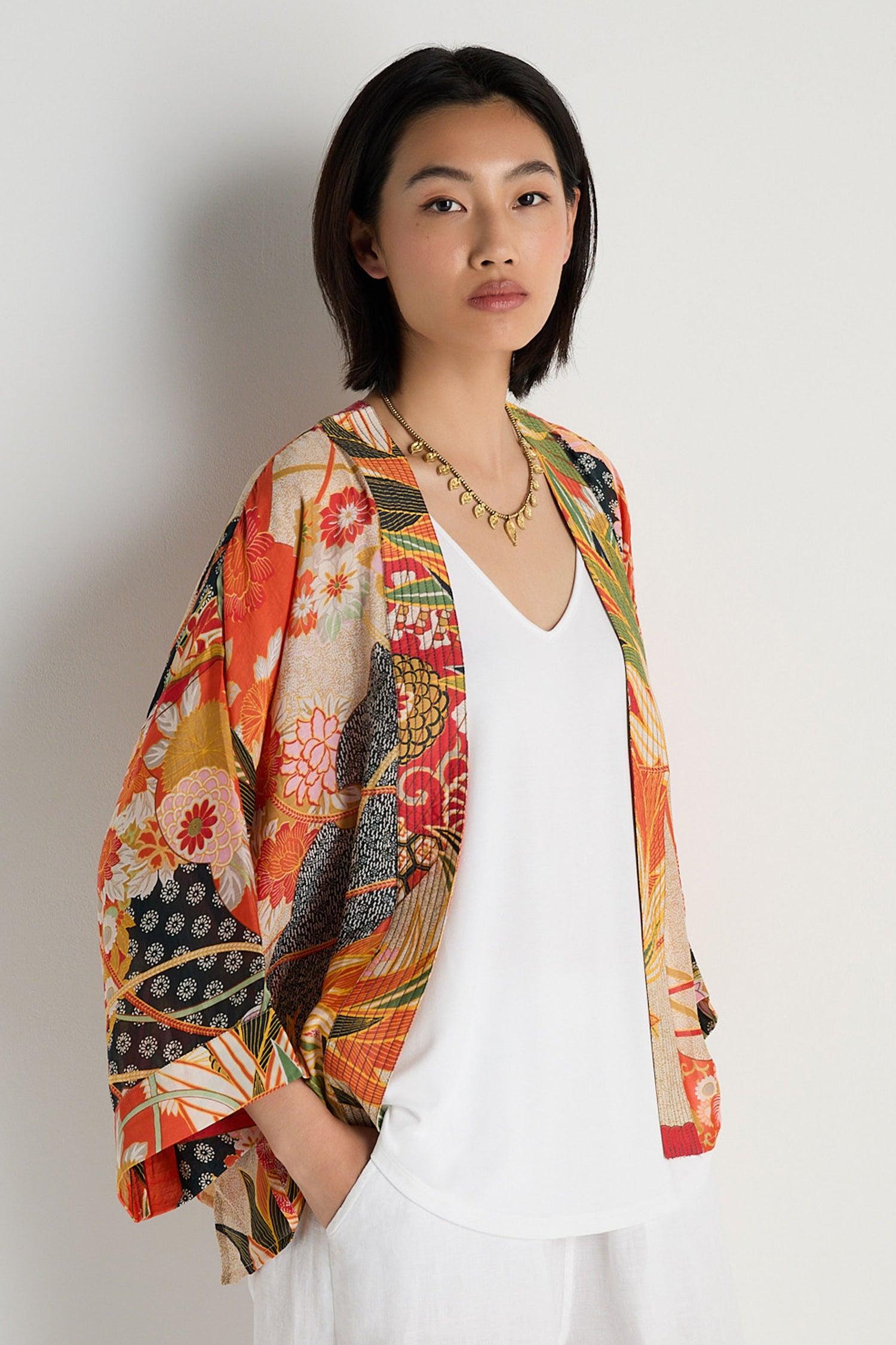 A woman stands against a white wall wearing the Fans Kimono, a colorful fan print jacket in a modal-cupro blend, layered over a white top and pants, with a gold necklace.