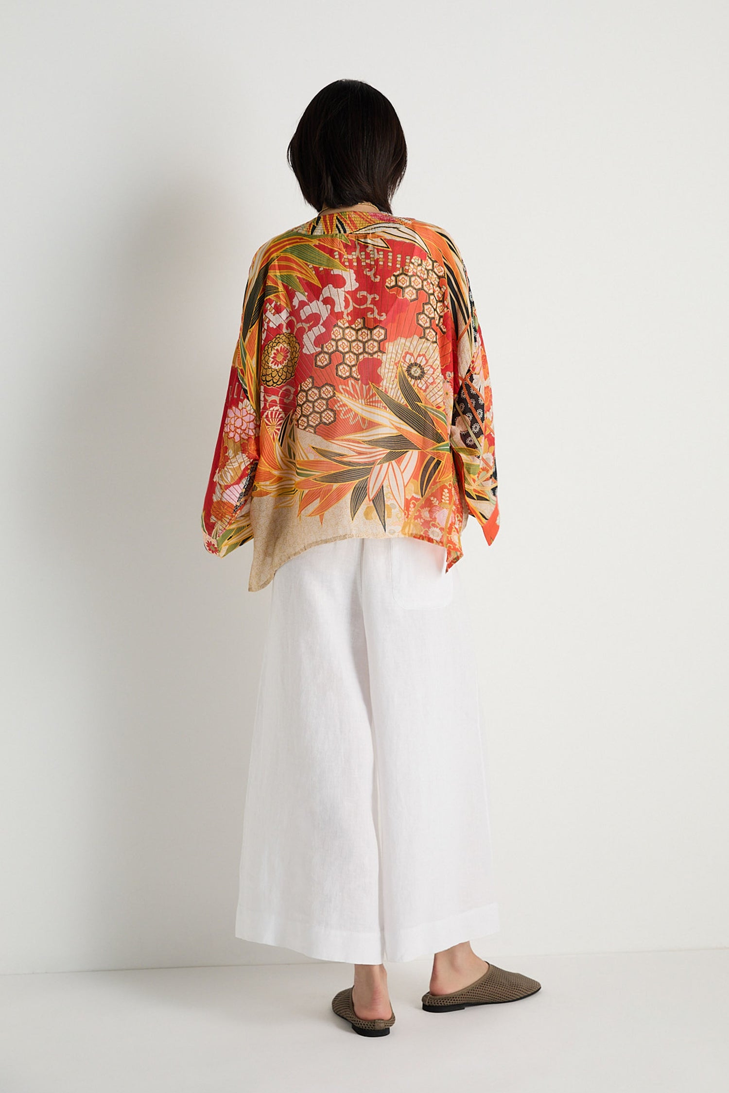 A person stands against a white wall, facing away, wearing the Fans Kimono—a vibrant fan print top in modal and cupro blend—paired with wide white pants and brown shoes.