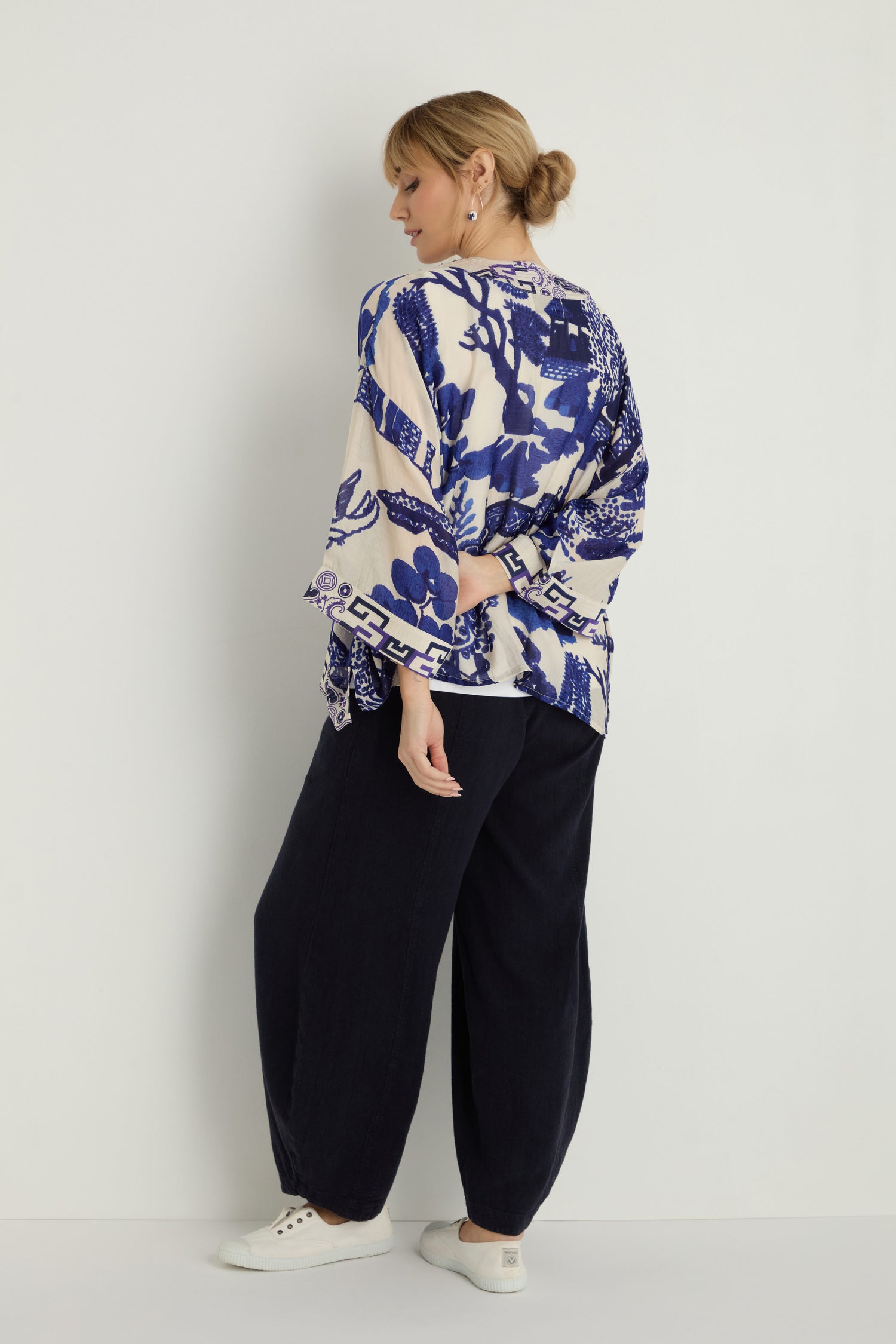 Facing a white wall, a person with light hair in a low bun wears the Willow Print Kimono, featuring bold blue-and-white willow motifs, paired with wide-leg black pants and white sneakers.