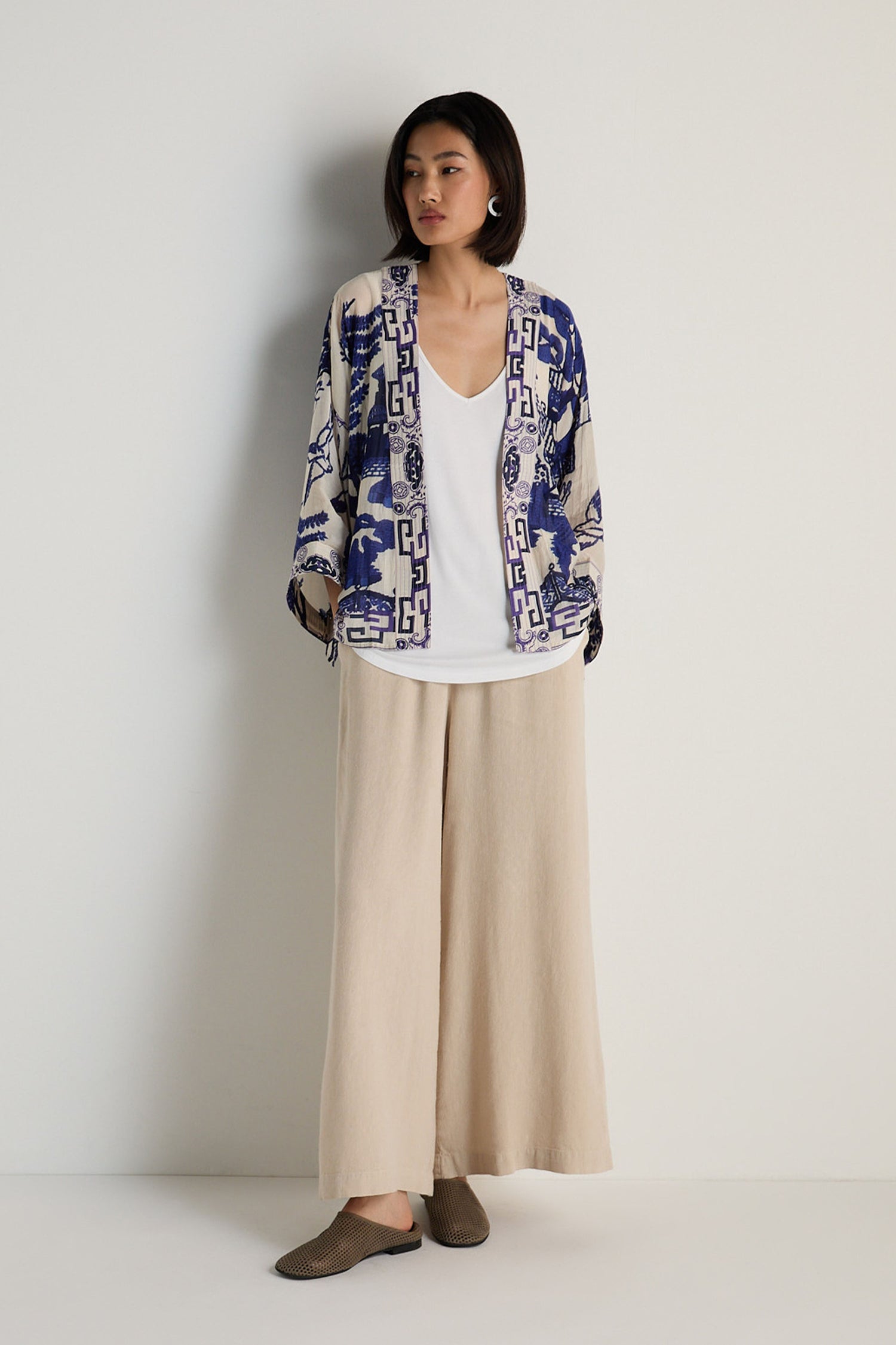 A woman stands against a white wall wearing the Willow Print Kimono, featuring a blue-and-white motif, paired with a white top, wide-leg beige pants, and brown shoes.