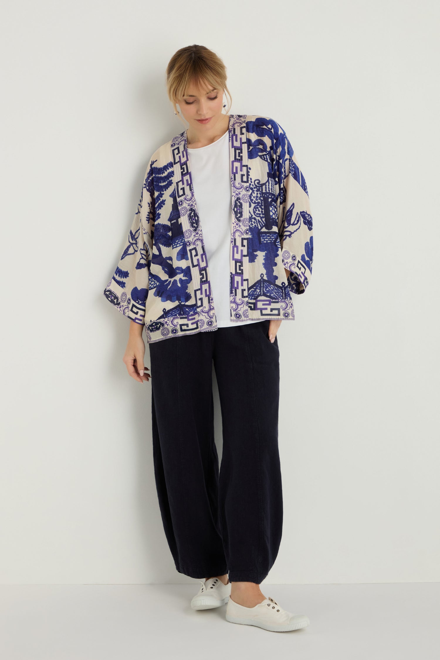 A woman stands against a white wall wearing the Willow Print Kimono over a white top, paired with loose black pants and white sneakers.