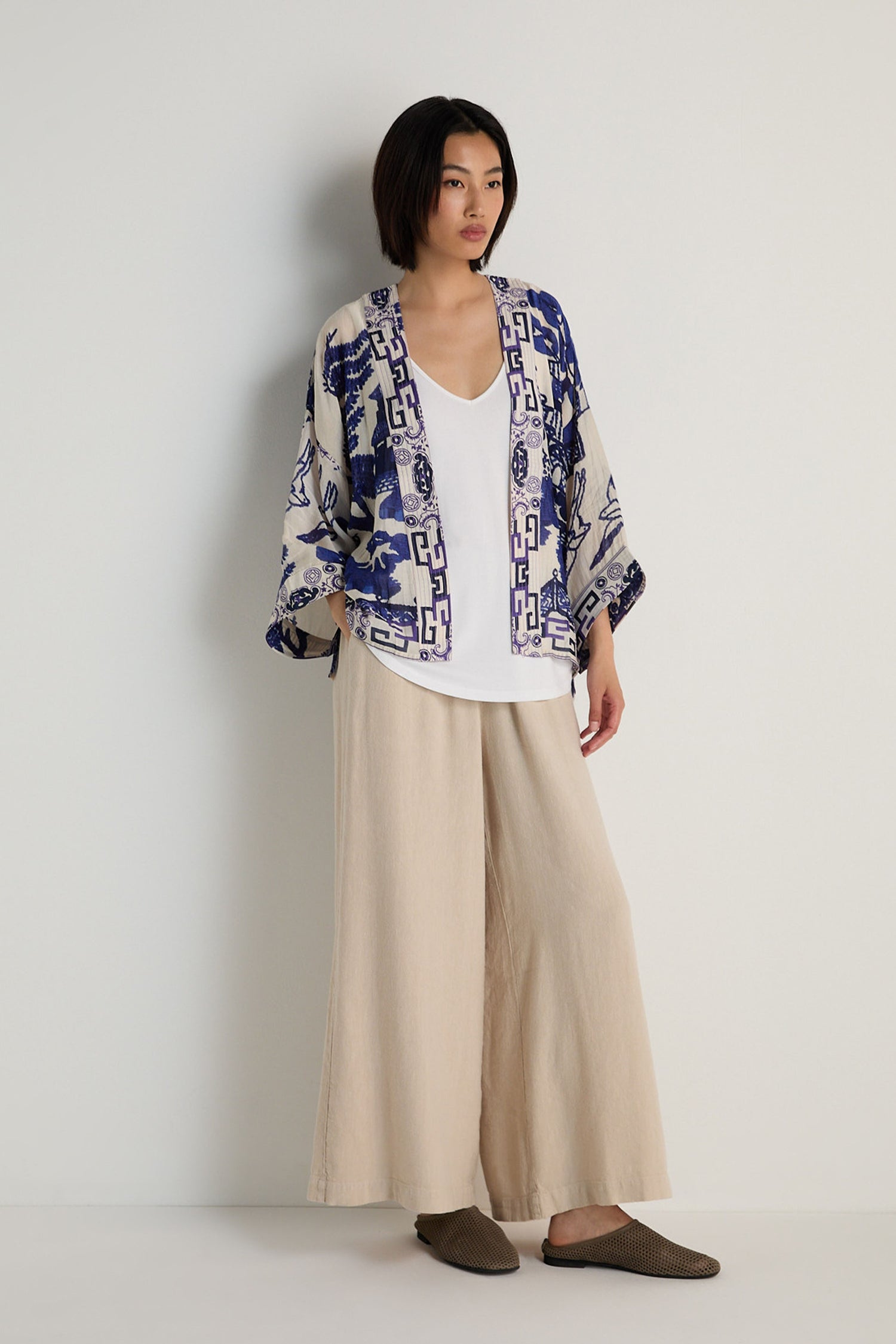 A woman stands against a plain wall wearing a white top, wide beige pants, and the Willow Print Kimono with a blue-and-white motif, styled with brown slippers for a relaxed look.