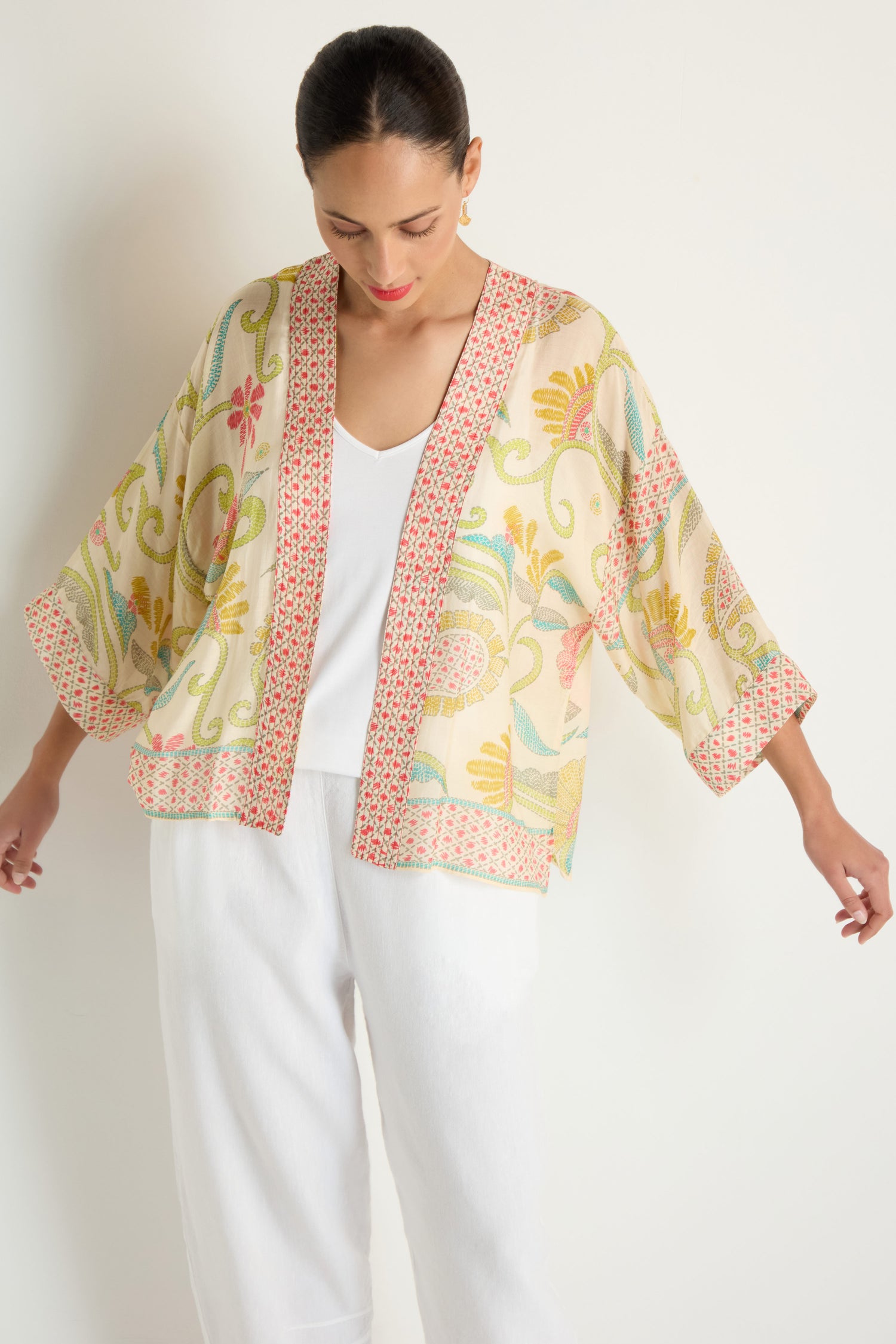 A woman models the Kantha Lime Kimono, featuring a lime-ecru palette and red trim in lightweight woven cloth, styled over a white top and pants against a plain background.