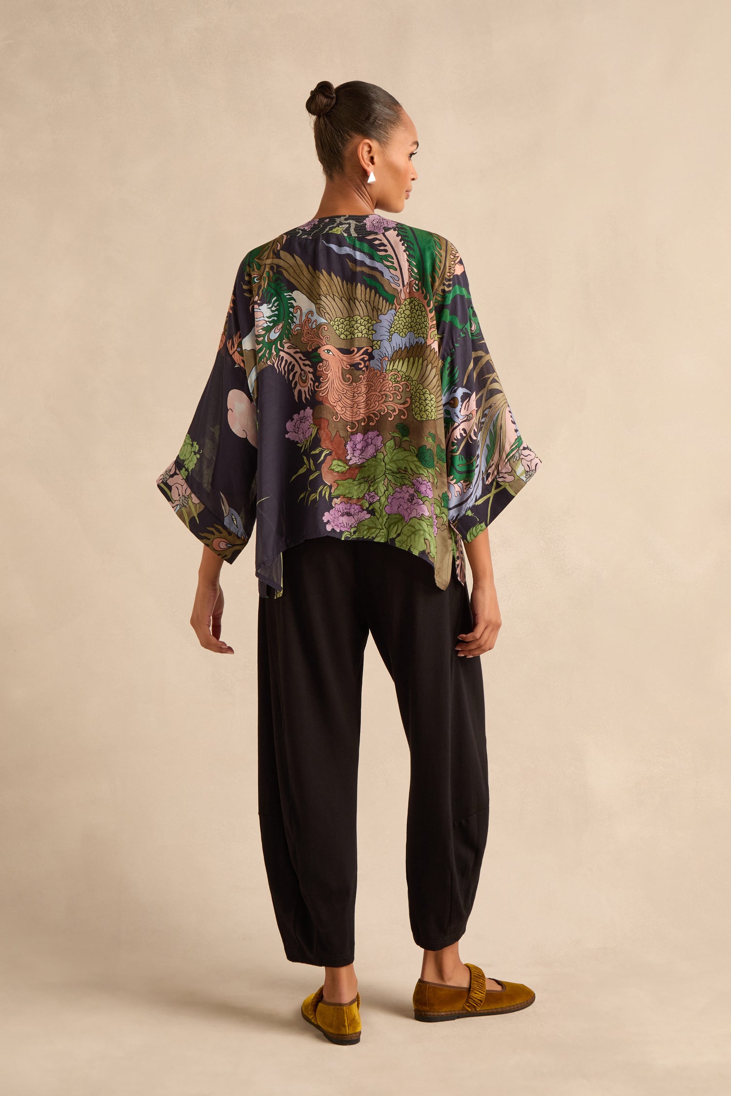 Person standing against a beige background, viewed from behind, wearing the loose floral Phoenix Kimono, black pants, and yellow shoes with hair in a bun.