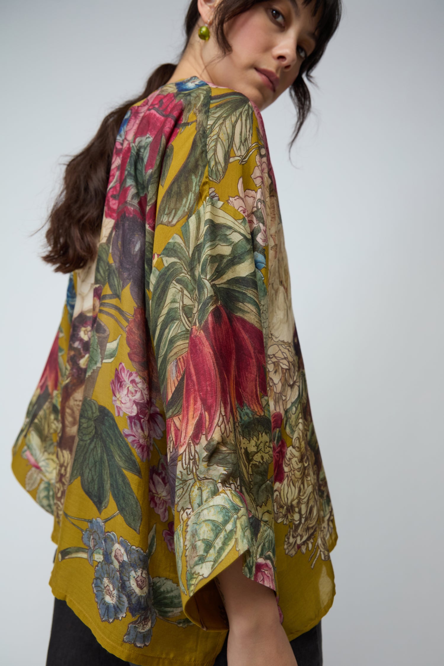 A woman with long dark hair poses against a plain light background, wearing the Primula Kimono—a lightweight piece featuring an artistic floral print.