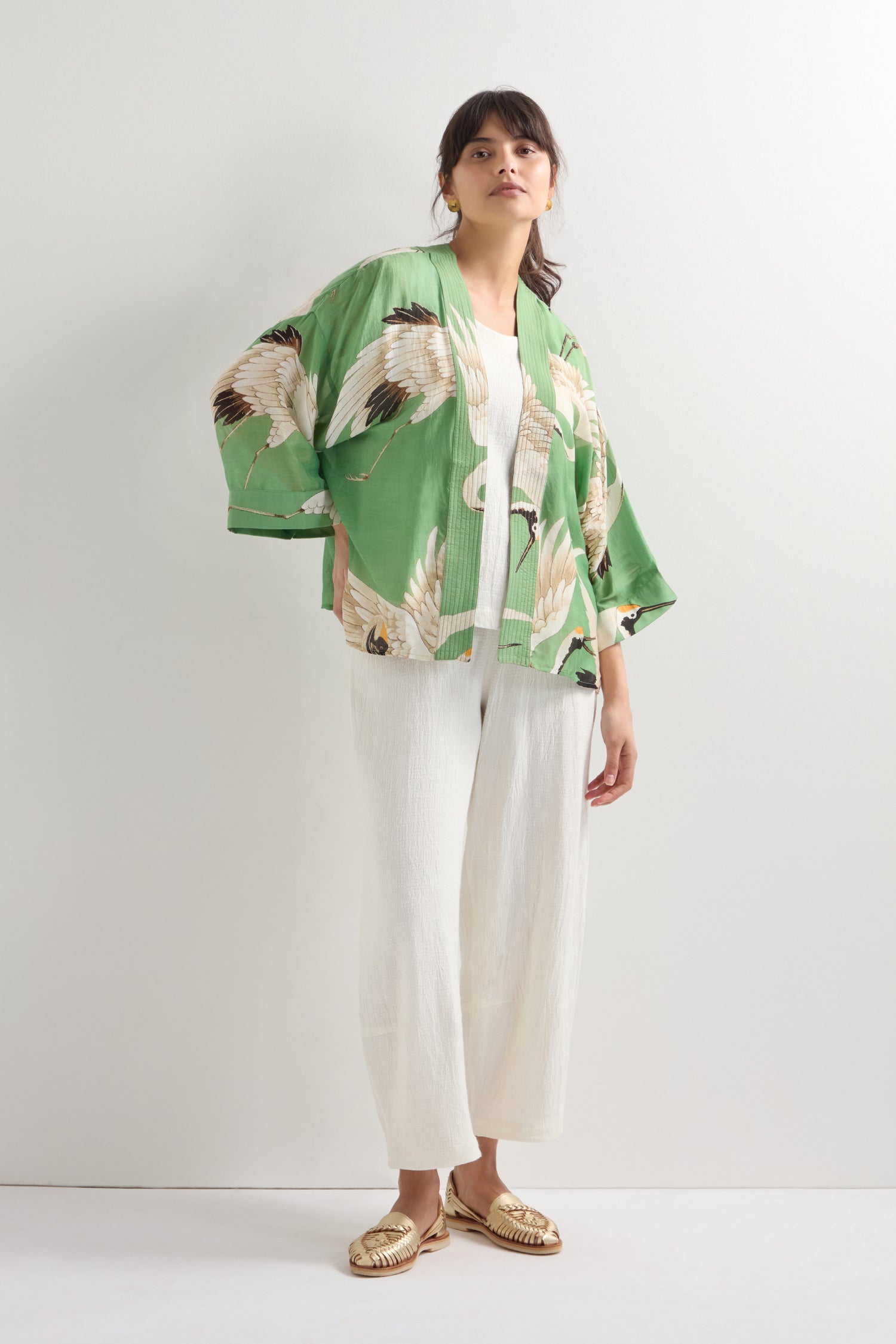 Someone is dressed in a Stork Print Kimono, along with a white top and pants, and gold sandals, standing against a plain light background.