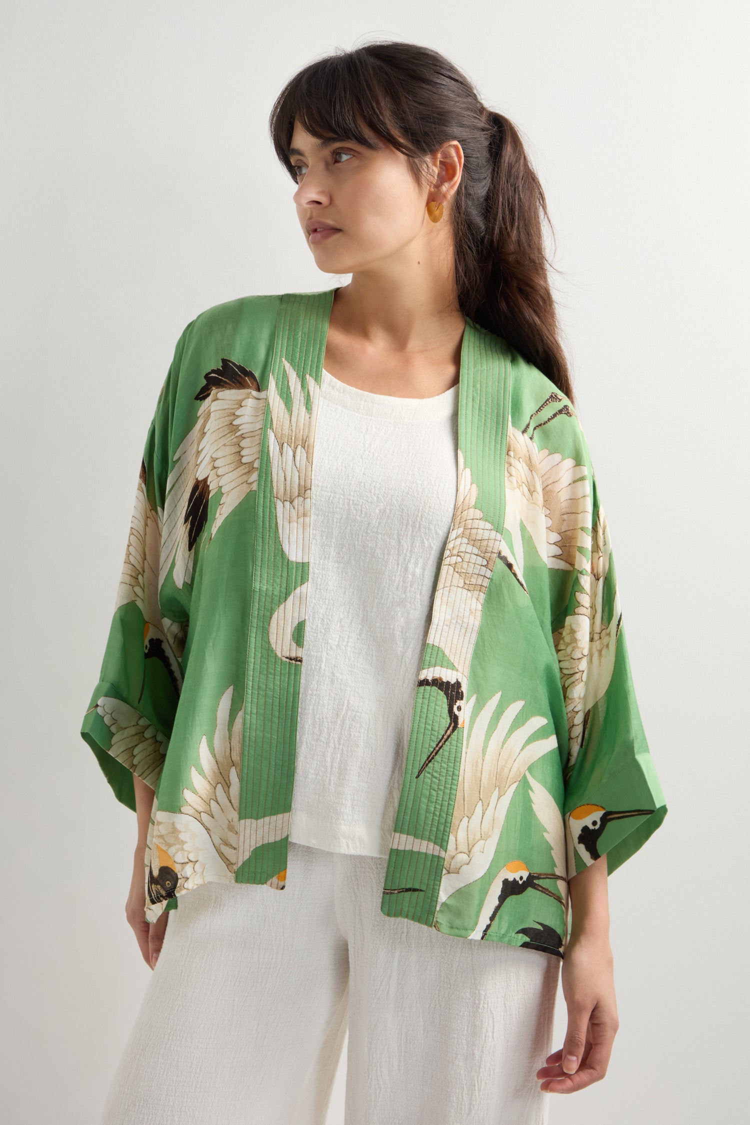 A woman stands wearing a "Stork Print Kimono," a green jacket with an Art Deco-inspired crane design, over a white top and pants, looking to the side.