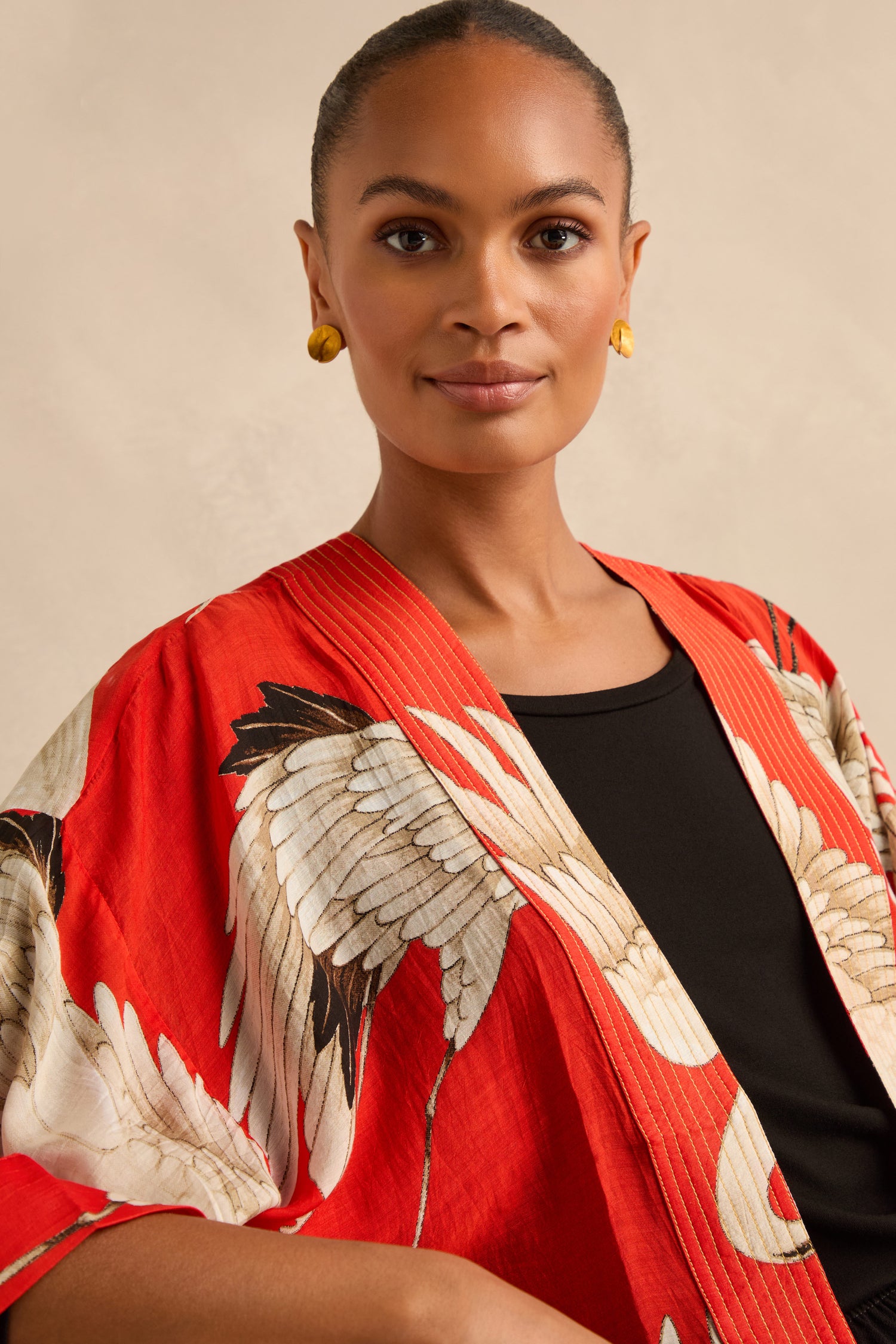 A woman with a neutral expression wears the Stork Kimono, featuring red floral patterns, over a black top and gold earrings against a light background.