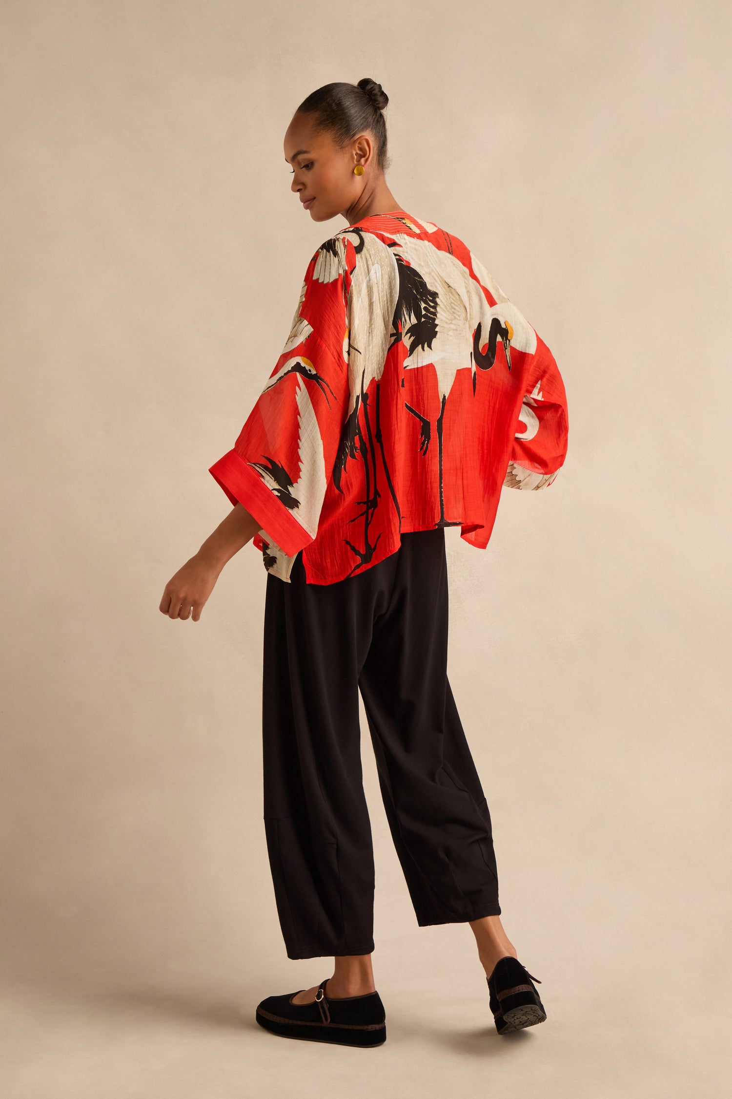 A person stands sideways wearing the Stork Kimono in a vibrant red, paired with black pants and black shoes against a plain beige background.