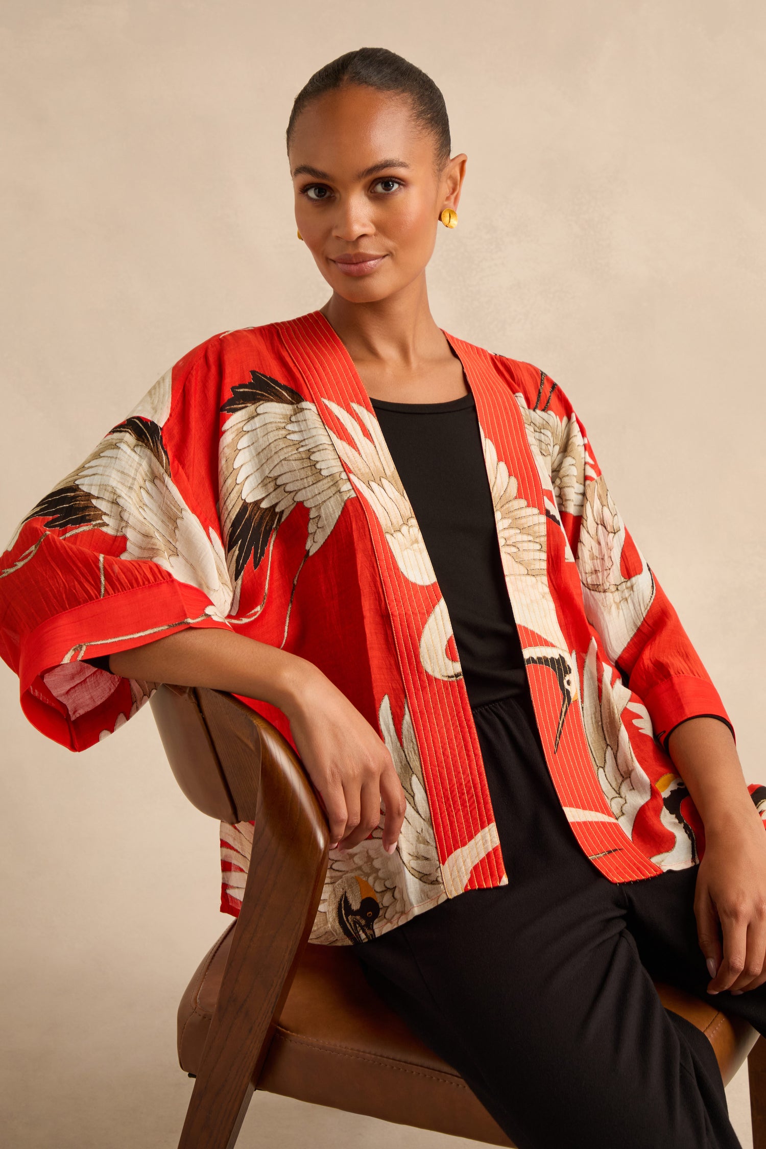 A woman seated on a brown chair against a neutral background wears the Stork Kimono, featuring a striking red and white print, layered over a black outfit.