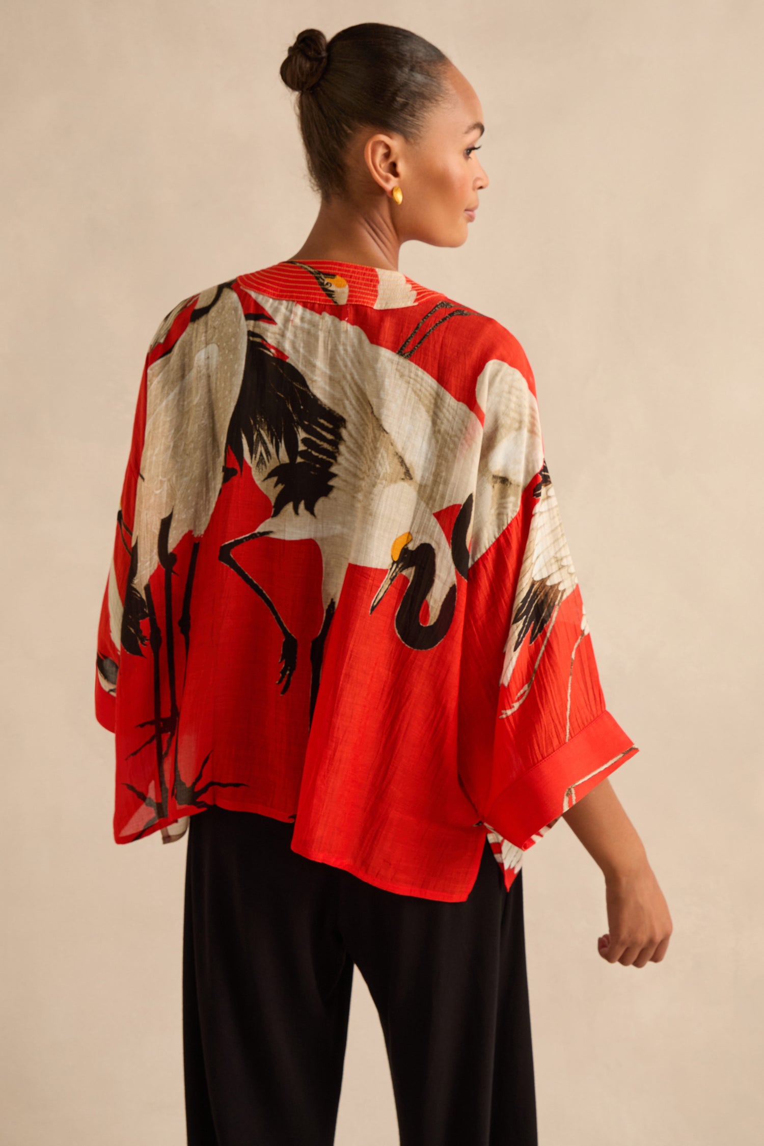 A woman models the Stork Kimono, a vibrant red Art Deco-inspired jacket featuring a bold white and black crane print, paired with black pants against a neutral background.