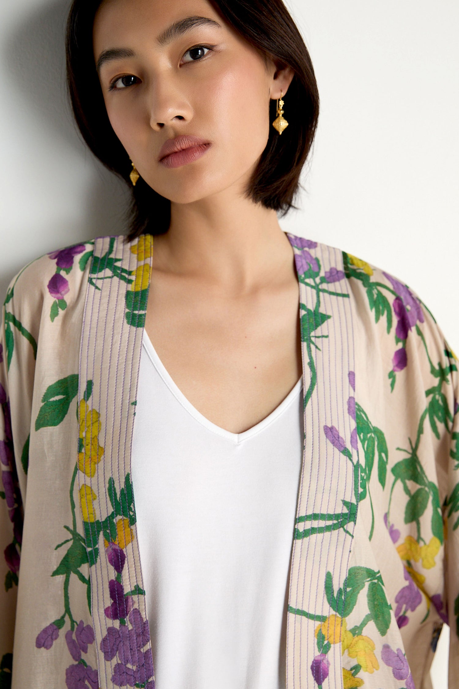 A woman wears the Wisteria Kimono, featuring a tan botanical print with purple and yellow flowers and green leaves, layered over a white V-neck shirt.