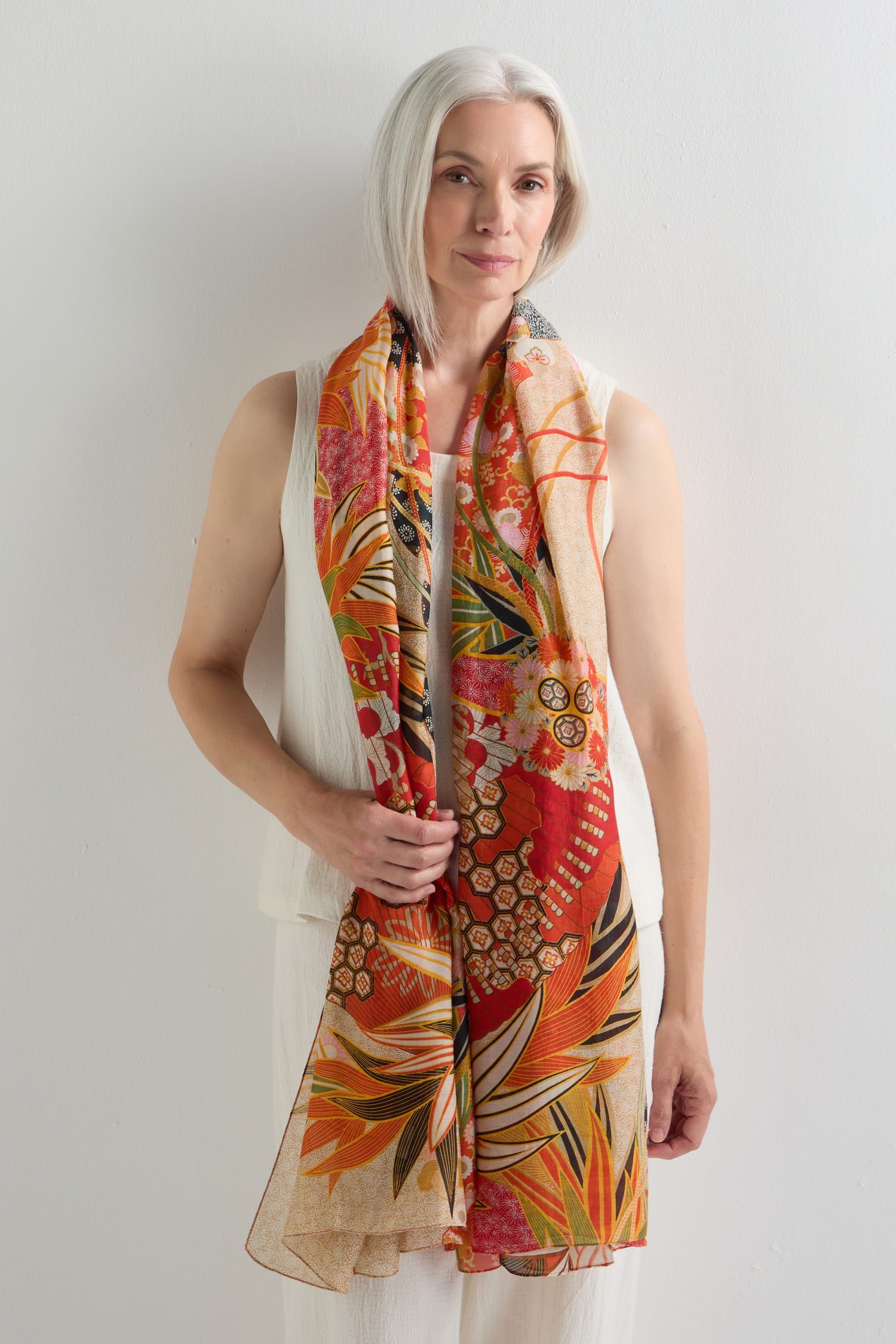 Woman with white hair in a sleeveless white top and pants poses against a plain white background, wearing the Fans Print Scarf draped stylishly around her neck.