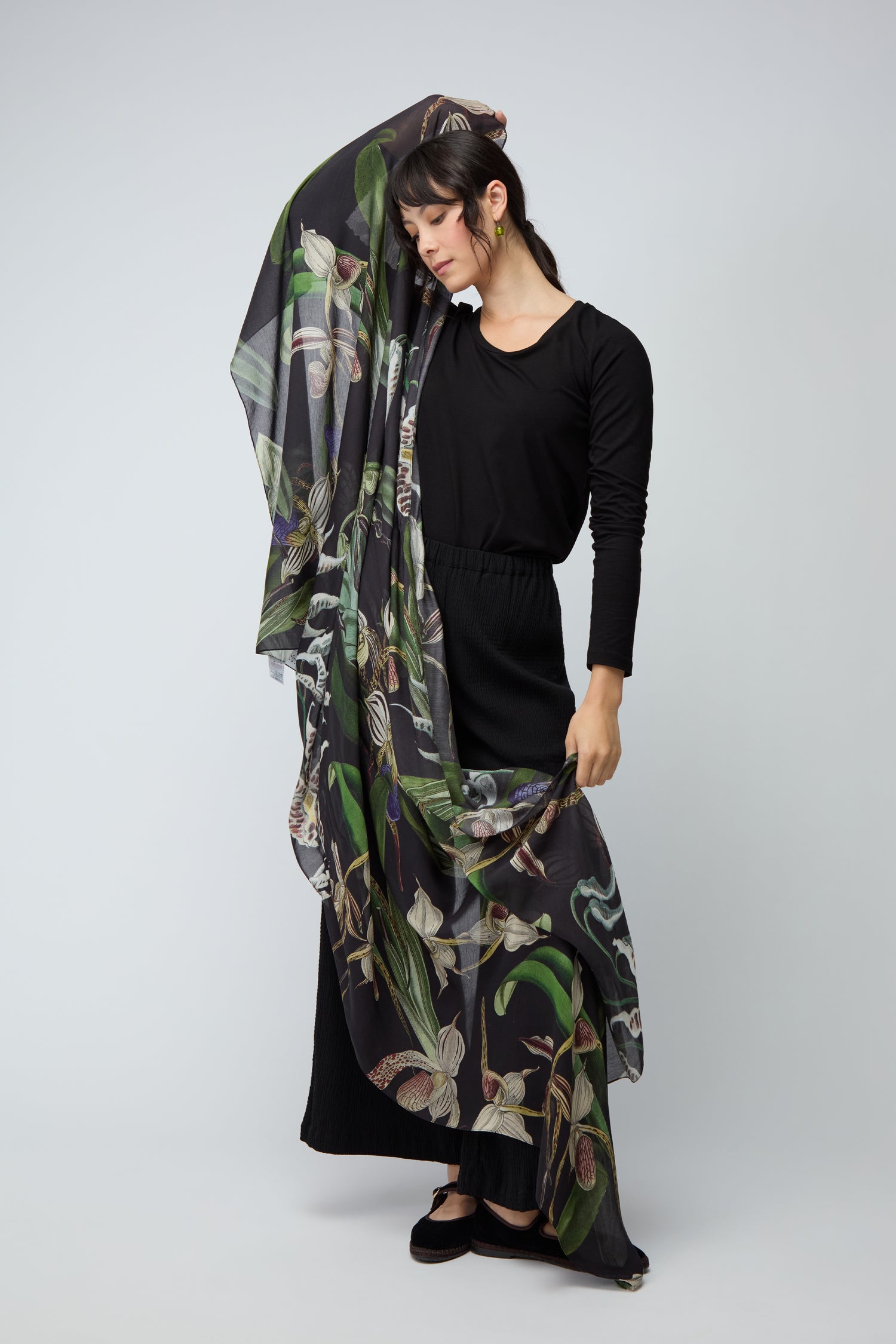 Wearing a black outfit, a woman holds up the Orchid Scarf, featuring a vibrant botanical print. Set against a plain light background, this elegant accessory enhances her ensemble with style and grace.