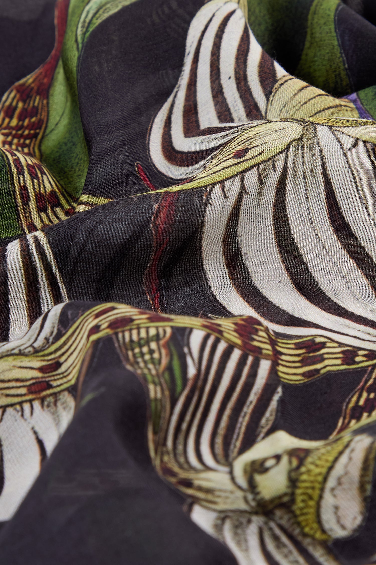 Close-up of the Orchid Scarf shows its botanical print with green and white striped flowers on a dark background, highlighting folds and texture—perfect as a versatile, elegant accessory.