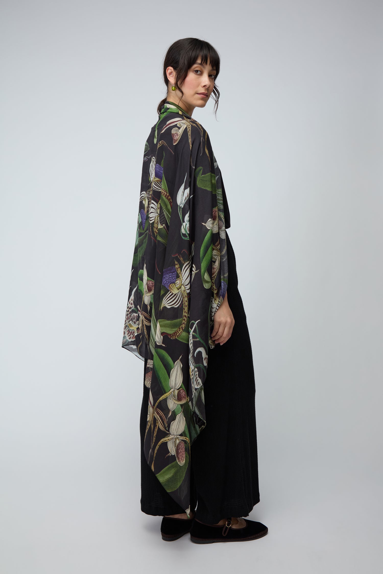 Standing sideways against a light gray background, a person wears the Orchid Scarf—a long, loose black garment with a vibrant botanical print—paired with black pants and black shoes.