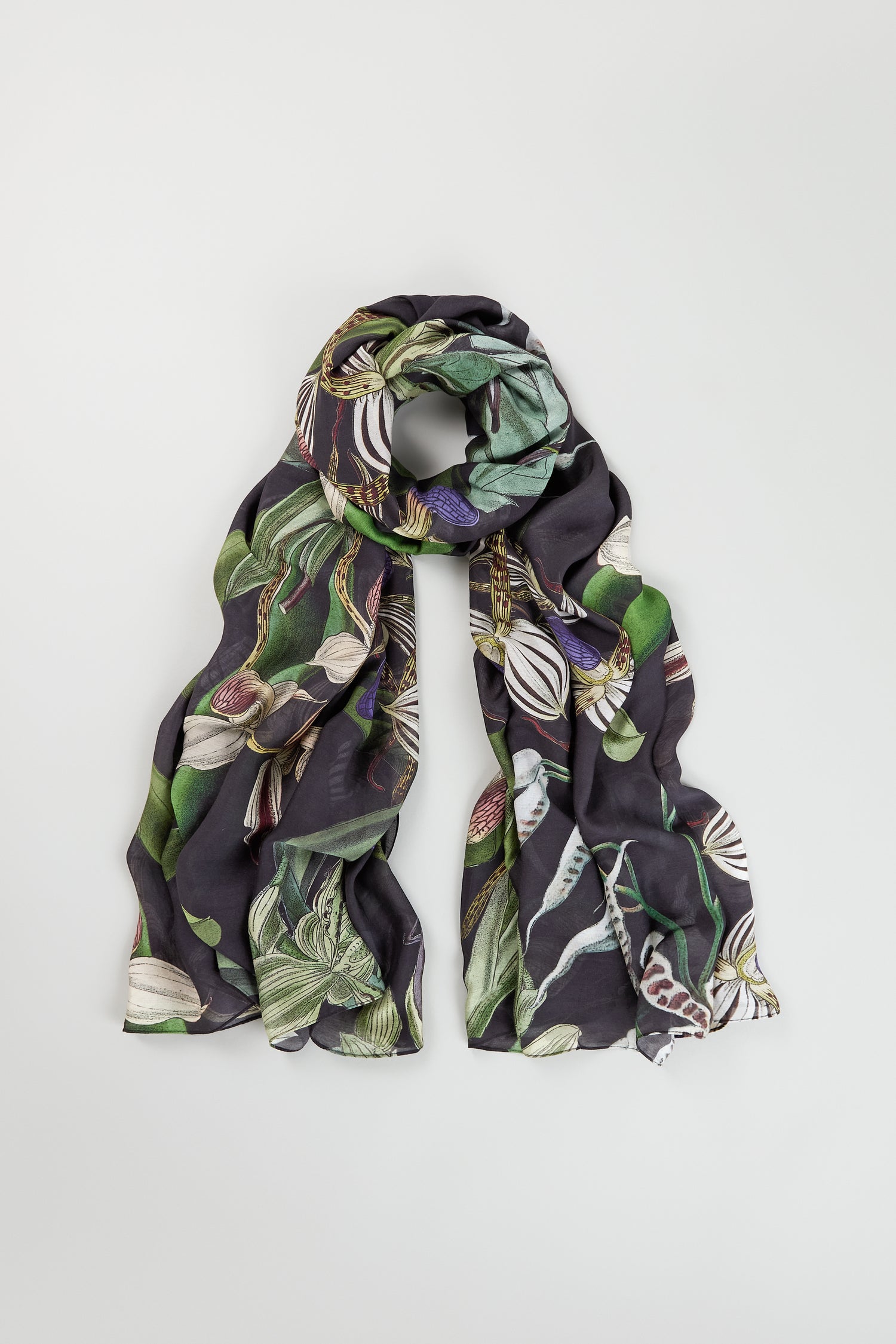 The Orchid Scarf features a dark base with green and white botanical prints, displayed in a loose loop on a light background—a stylish and versatile accessory for any wardrobe.