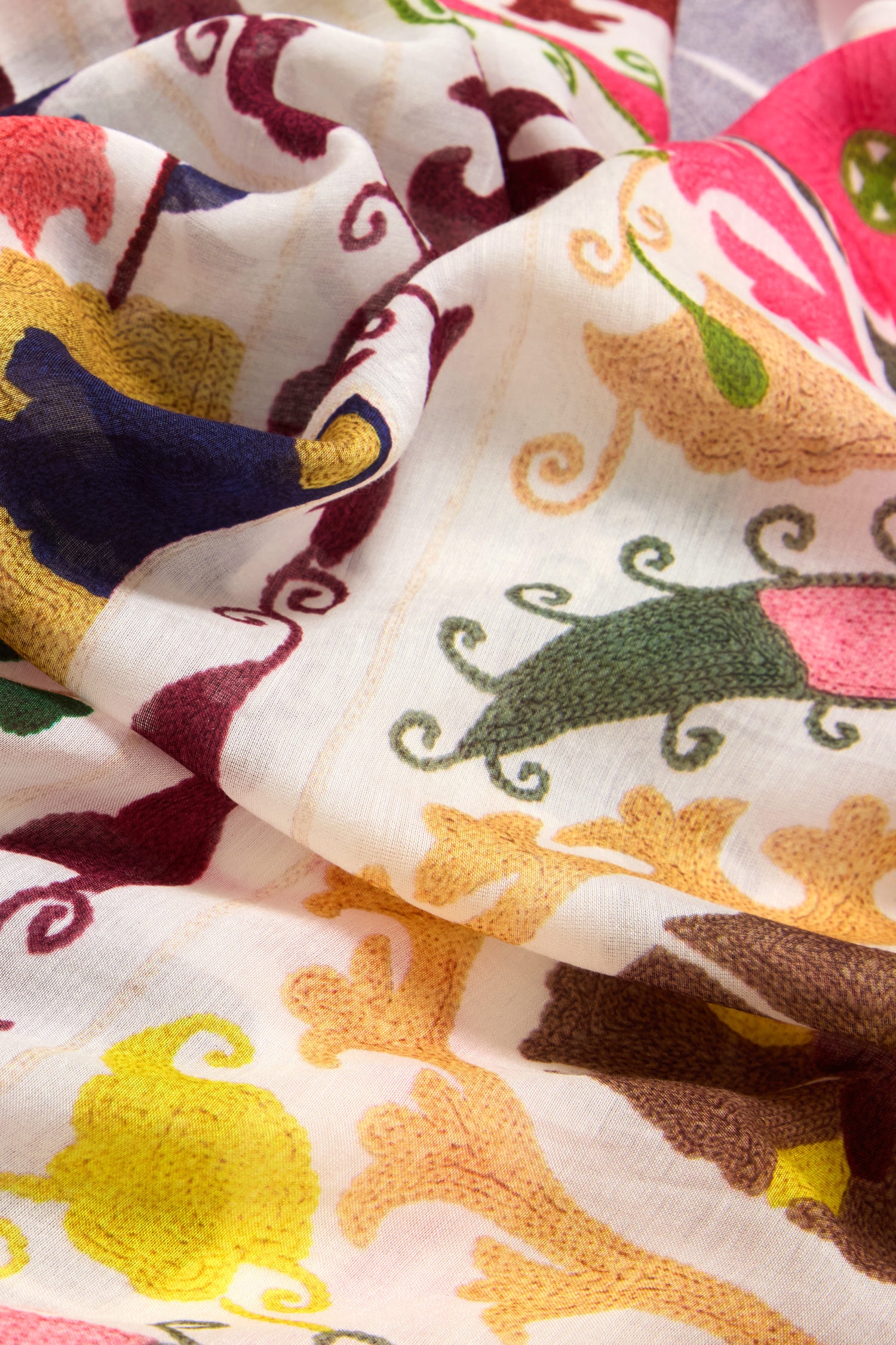 The Suzani Print Scarf showcases a versatile design with colorful fabric featuring intricate swirling needlework in red, yellow, green, and blue on a white background.