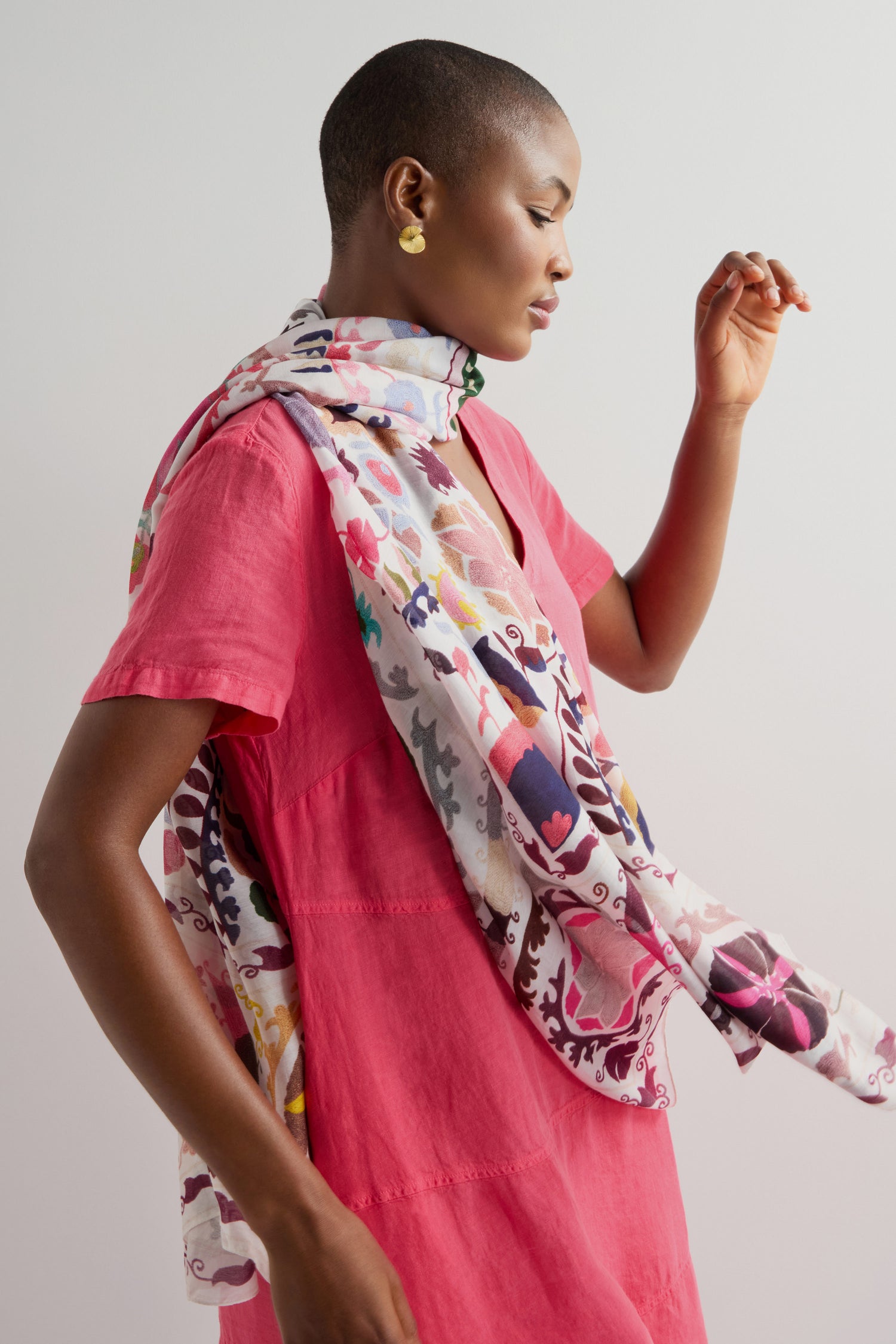 A person in a pink dress, wearing a versatile Suzani Print Scarf with intricate floral needlework, stands against a plain background.