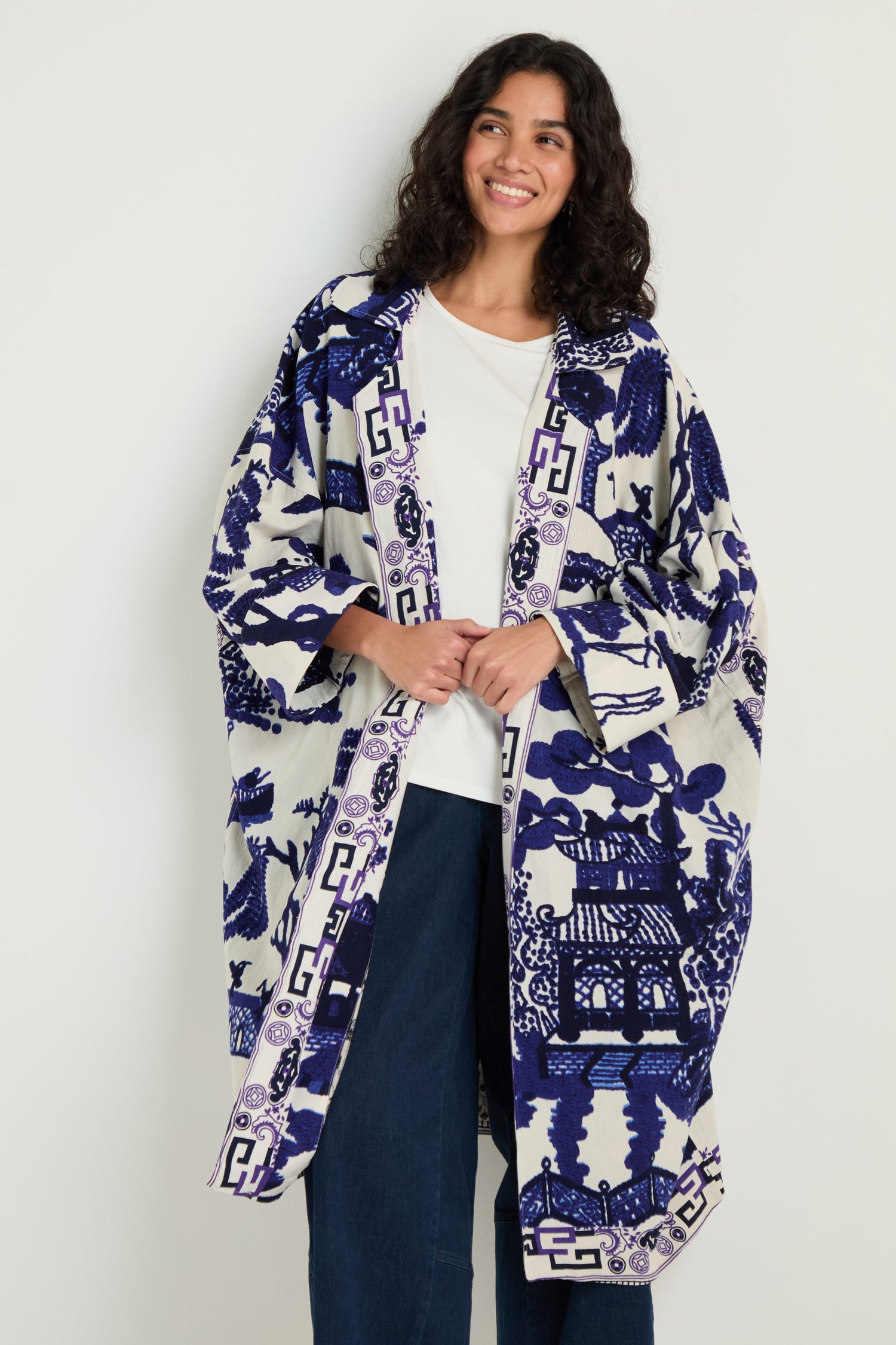 A person wearing a white shirt, blue jeans, and the Willow Print Summer Coat—a blue and white patterned summer layer—stands smiling against a plain light background.