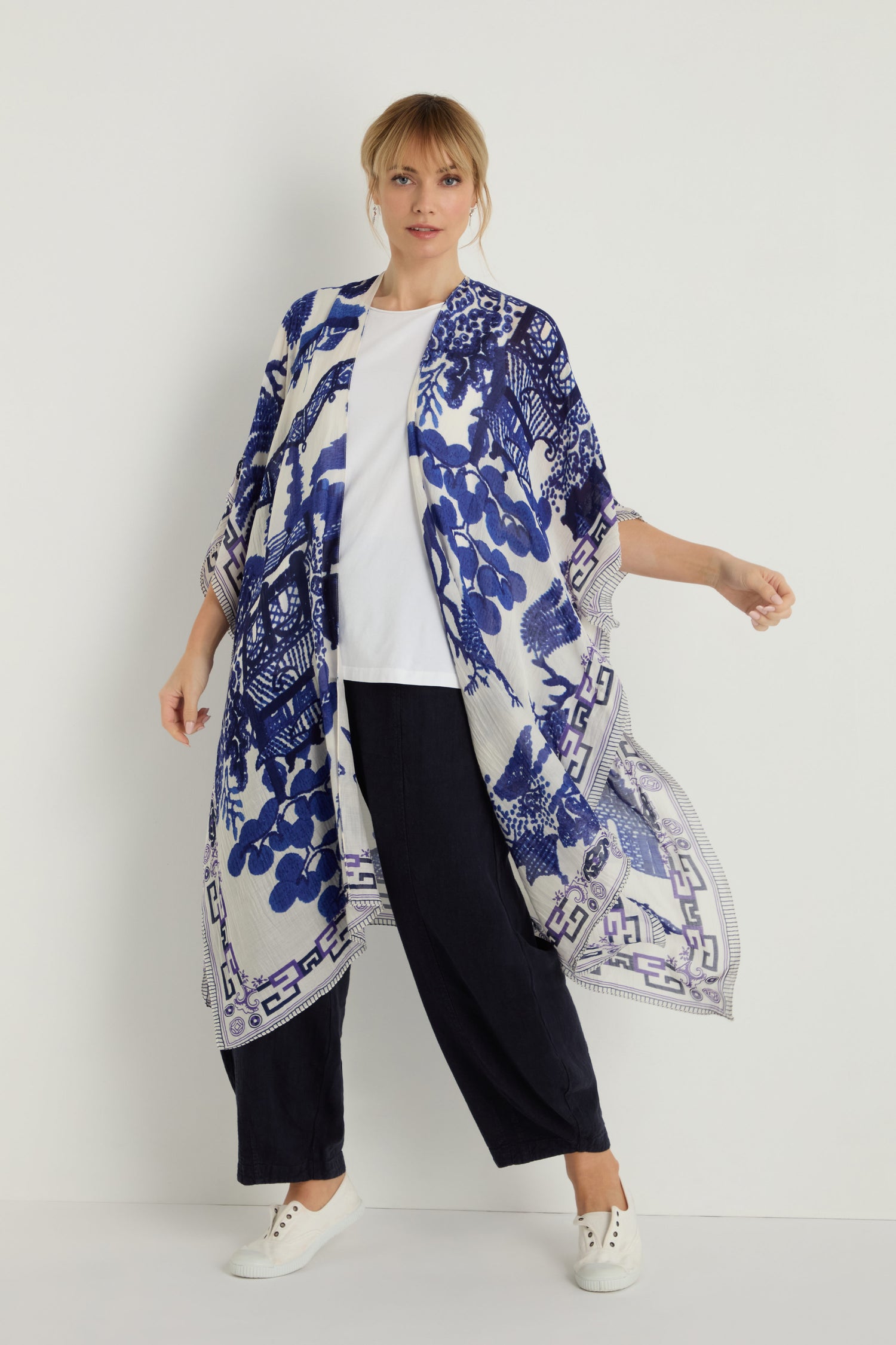 A woman stands against a white background wearing the Willow Print Throwover—a blue and white patterned kimono made from sustainable fabric—layered over a white top, black pants, and white sneakers.
