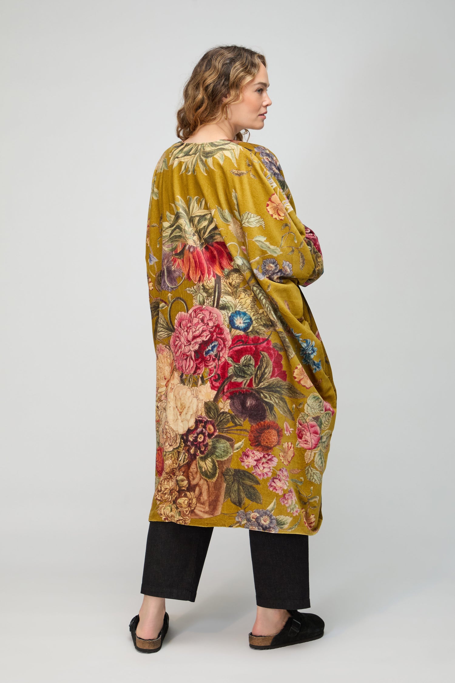 A woman faces away in a long mustard-yellow Primula Velvet Grande Kimono, featuring a colorful floral pattern, worn with black pants and shoes against a plain background.