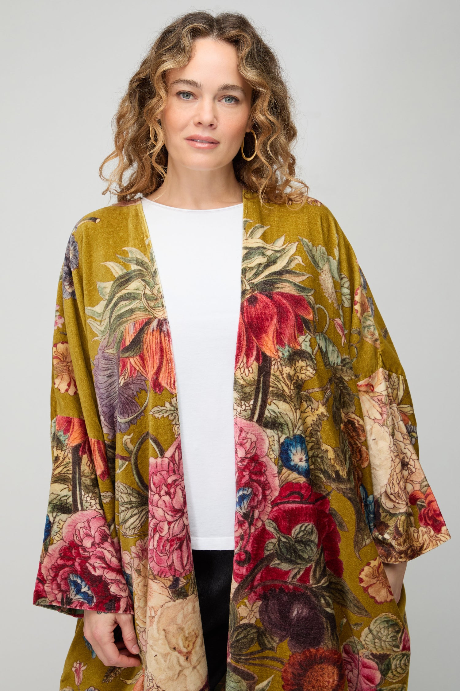 A woman with curly hair wears a white shirt and the Primula Velvet Grande Kimono by One Hundred Stars, featuring a long, floral pattern, standing against a plain light gray background.