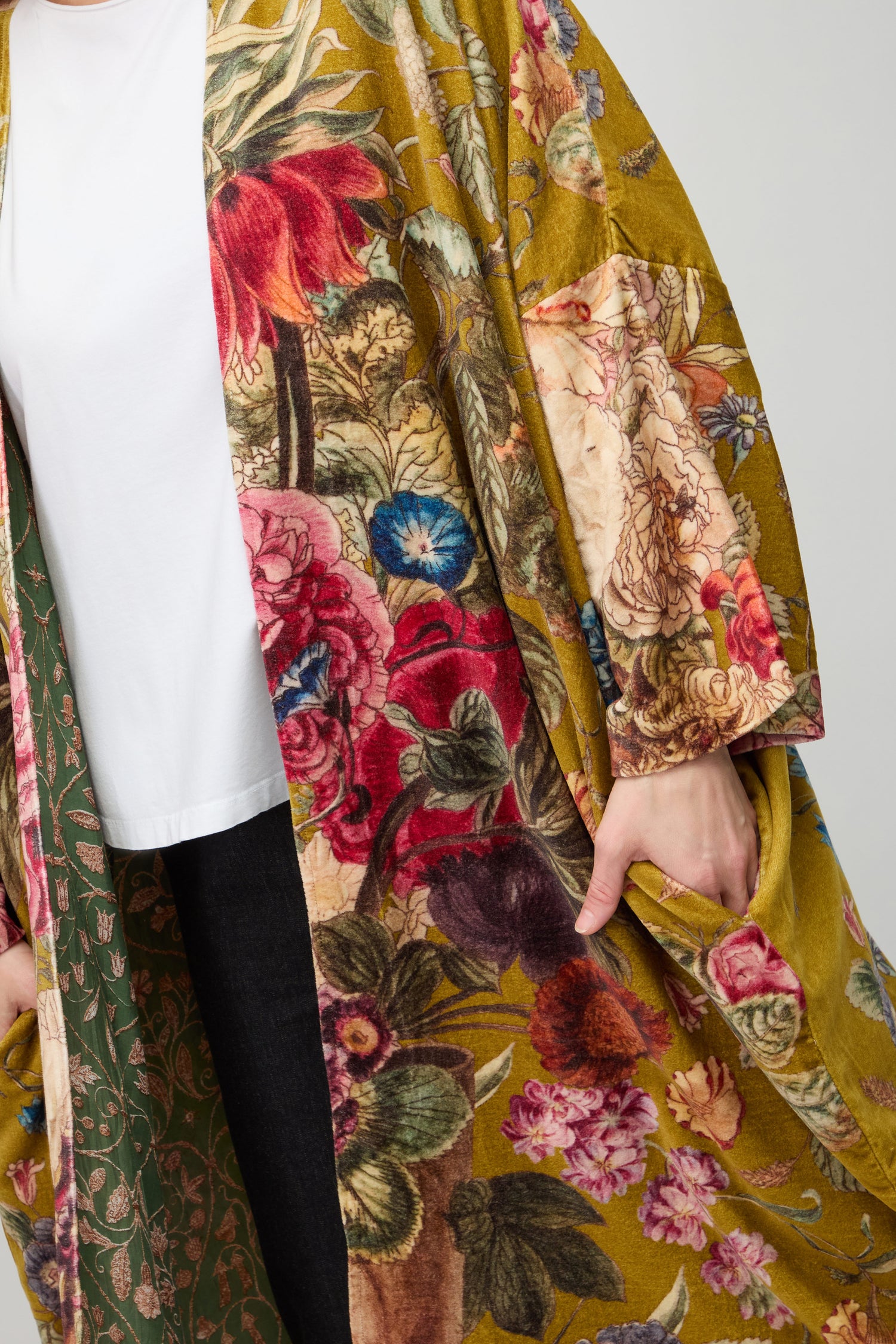 A person wears a white shirt, dark pants, and the Primula Velvet Grande Kimono—long, colorful, floral-patterned silk velvet—with one hand in their pocket.