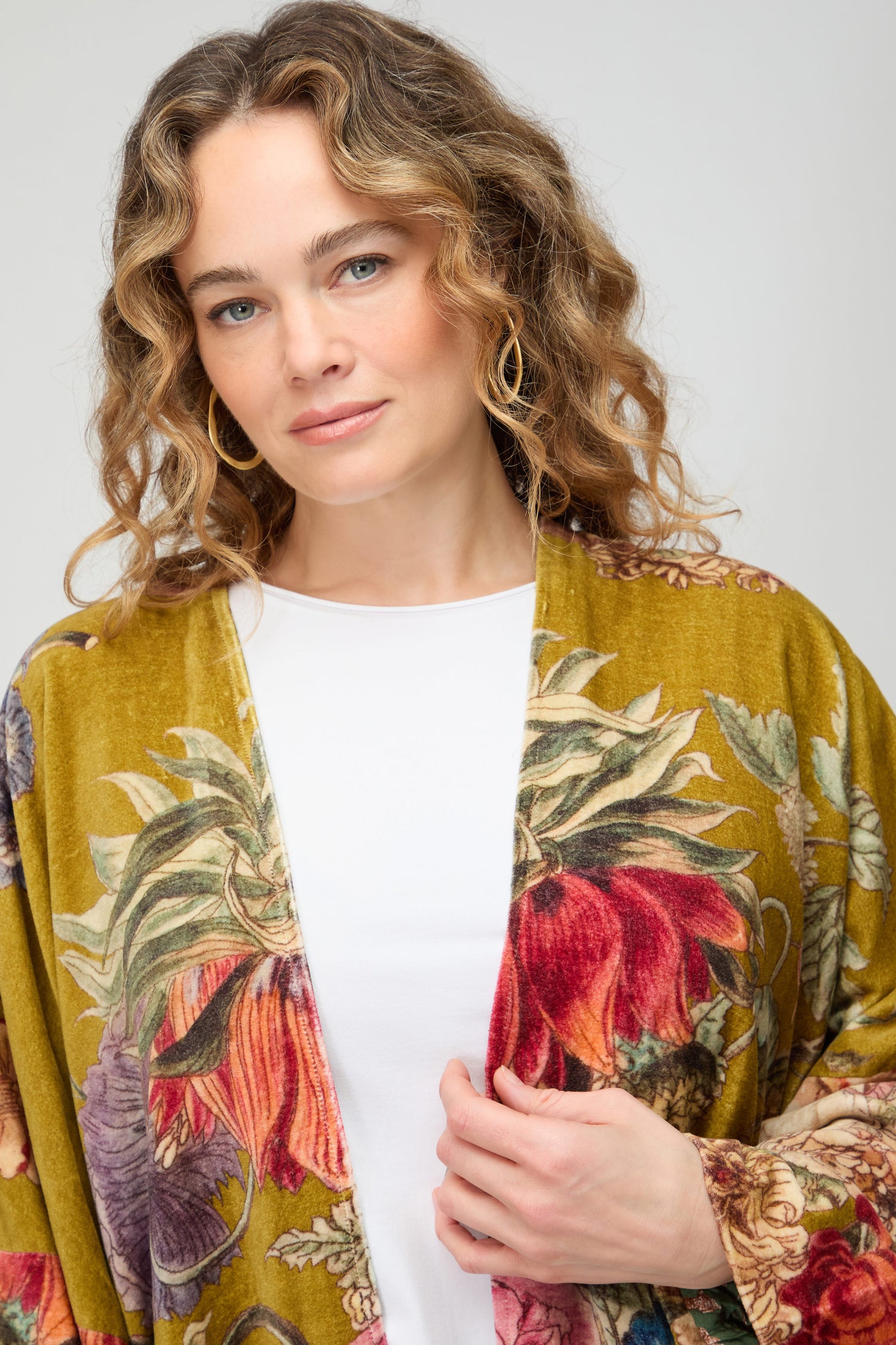 A woman with wavy hair wears the Primula Velvet Grande Kimono—a floral-patterned yellow-green silk velvet kimono—over a white shirt, standing against a plain background.