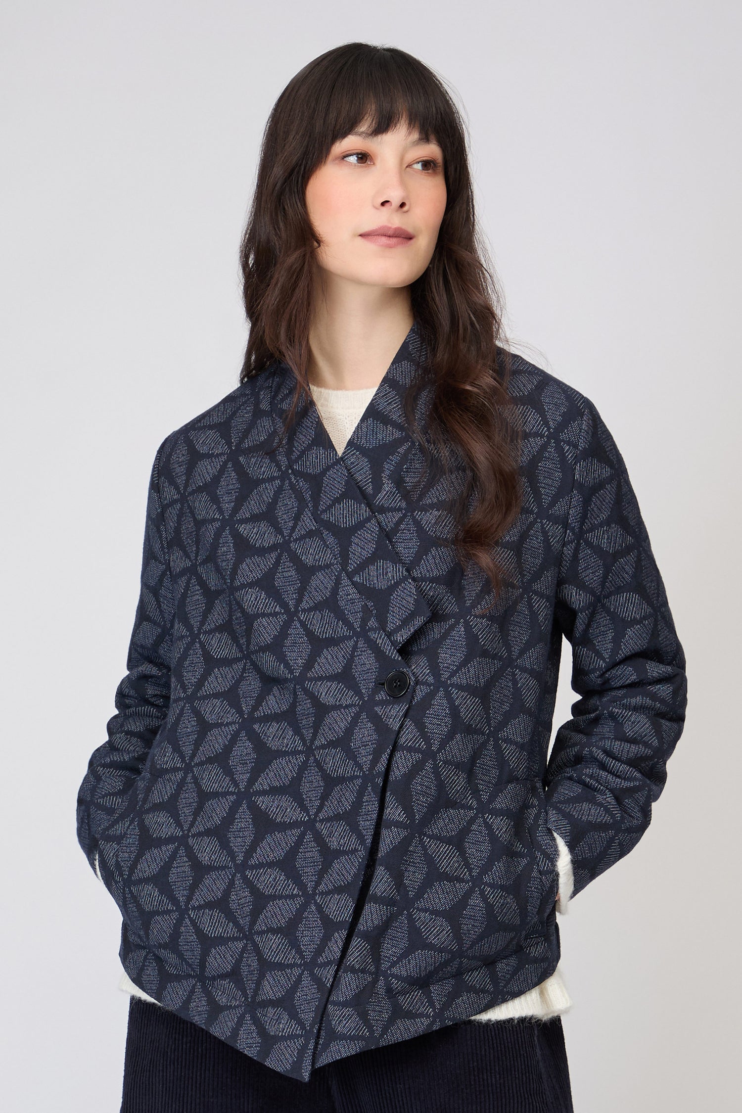A woman stands against a plain light background, wearing the Diamond Floral Indigo Jacket with a geometric pattern and two buttons over matching dark pants.