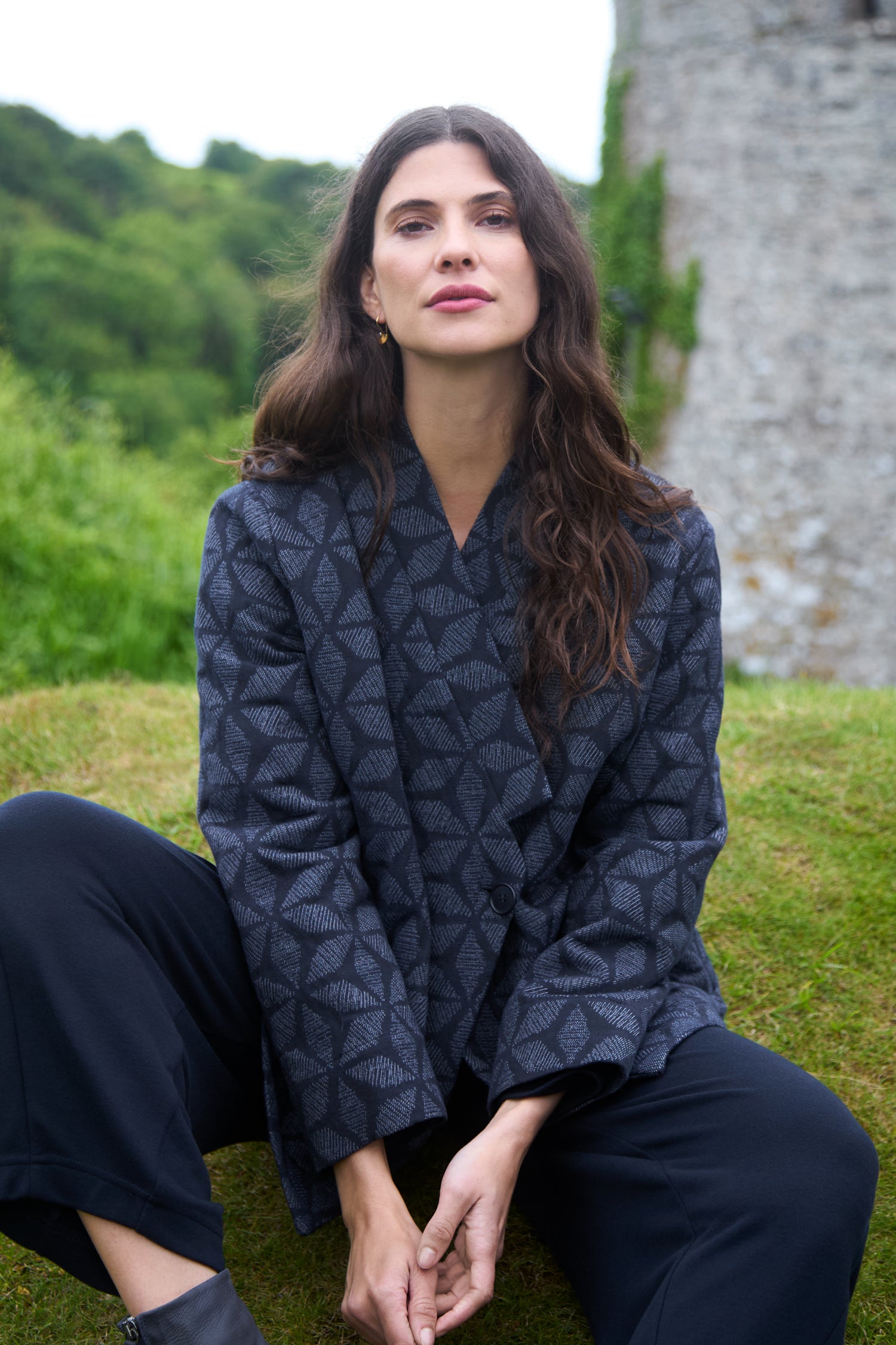 A woman with long brown hair sits on the grass wearing a Diamond Floral Indigo Jacket and black pants, with a stone structure and greenery in the background.
