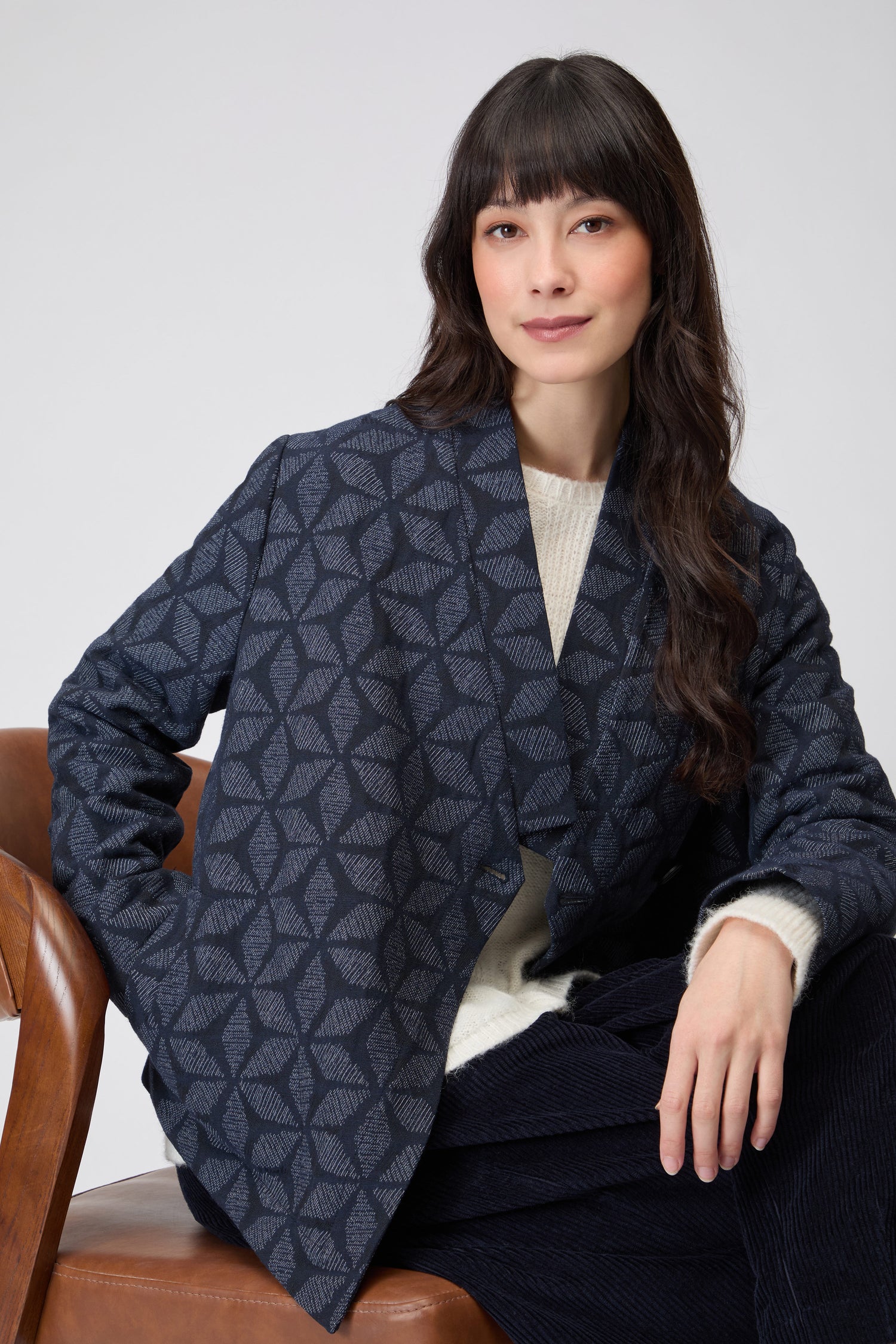 A woman in a Diamond Floral Indigo Jacket sits on a wooden stool against a plain background.