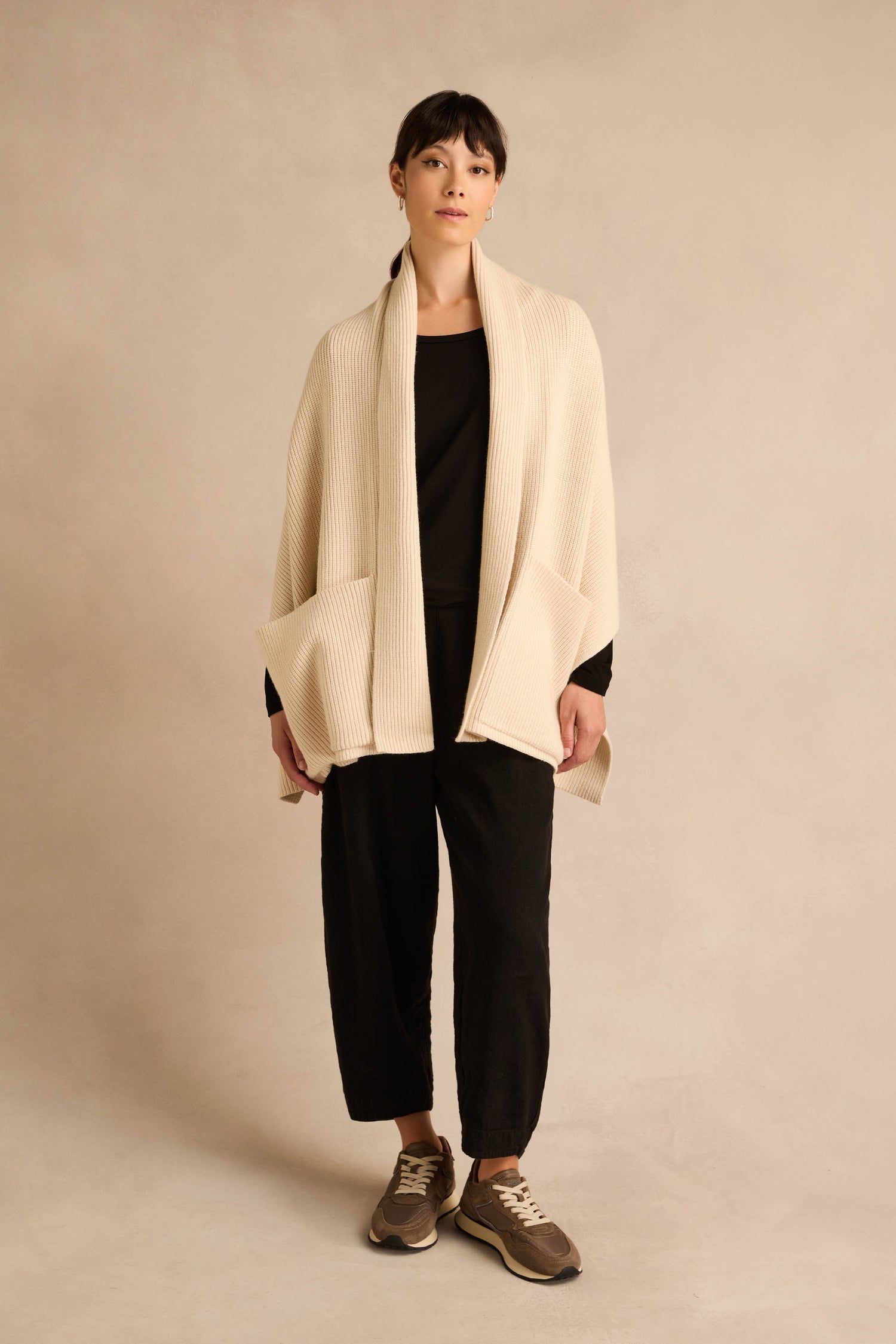 A person stands against a neutral background wearing a black outfit, accessorized with the Cream Pocket Scarf—crafted from a recycled fiber blend with ribbed detailing—and finished with brown sneakers.