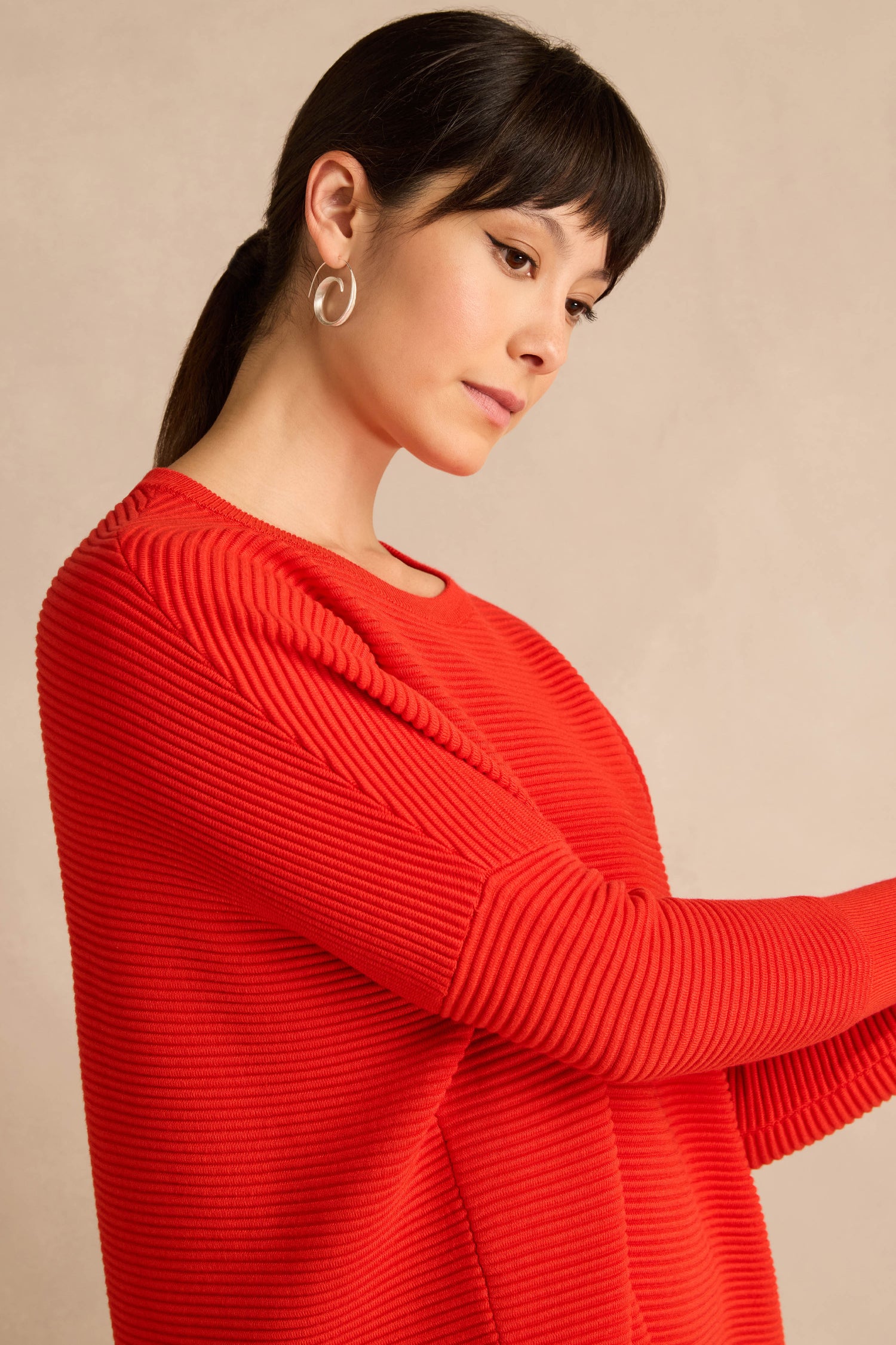 A woman with straight dark hair styled in a ponytail wears the Red Ribbed Jumper, paired with silver hoop earrings, as she poses against a plain beige background.
