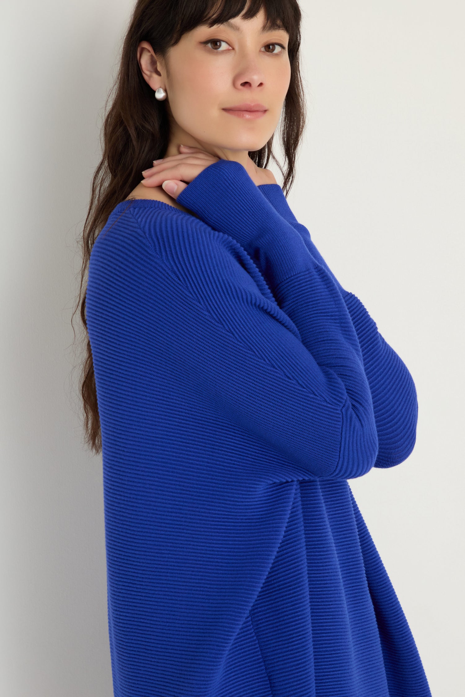 A woman with long dark hair, wearing pearl earrings and a Paisie Round Neck Ribbed Top in royal blue, stands against a white background with her hands gently crossed.