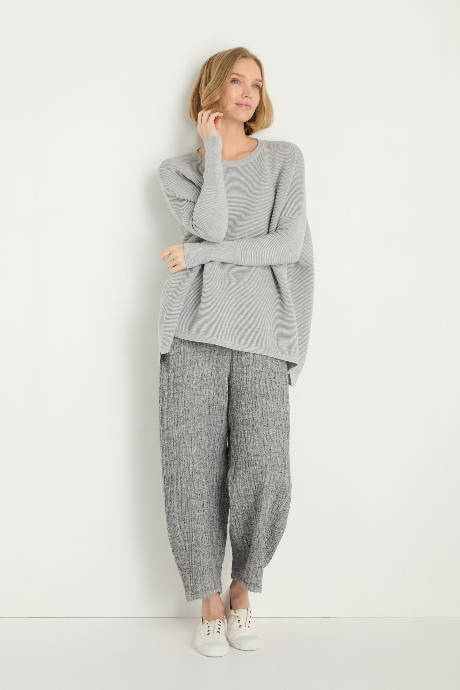 A woman stands against a white wall wearing a Round Neck Ribbed Top in grey, textured grey pants, and white sneakers, gently touching her chin with a slight smile.
