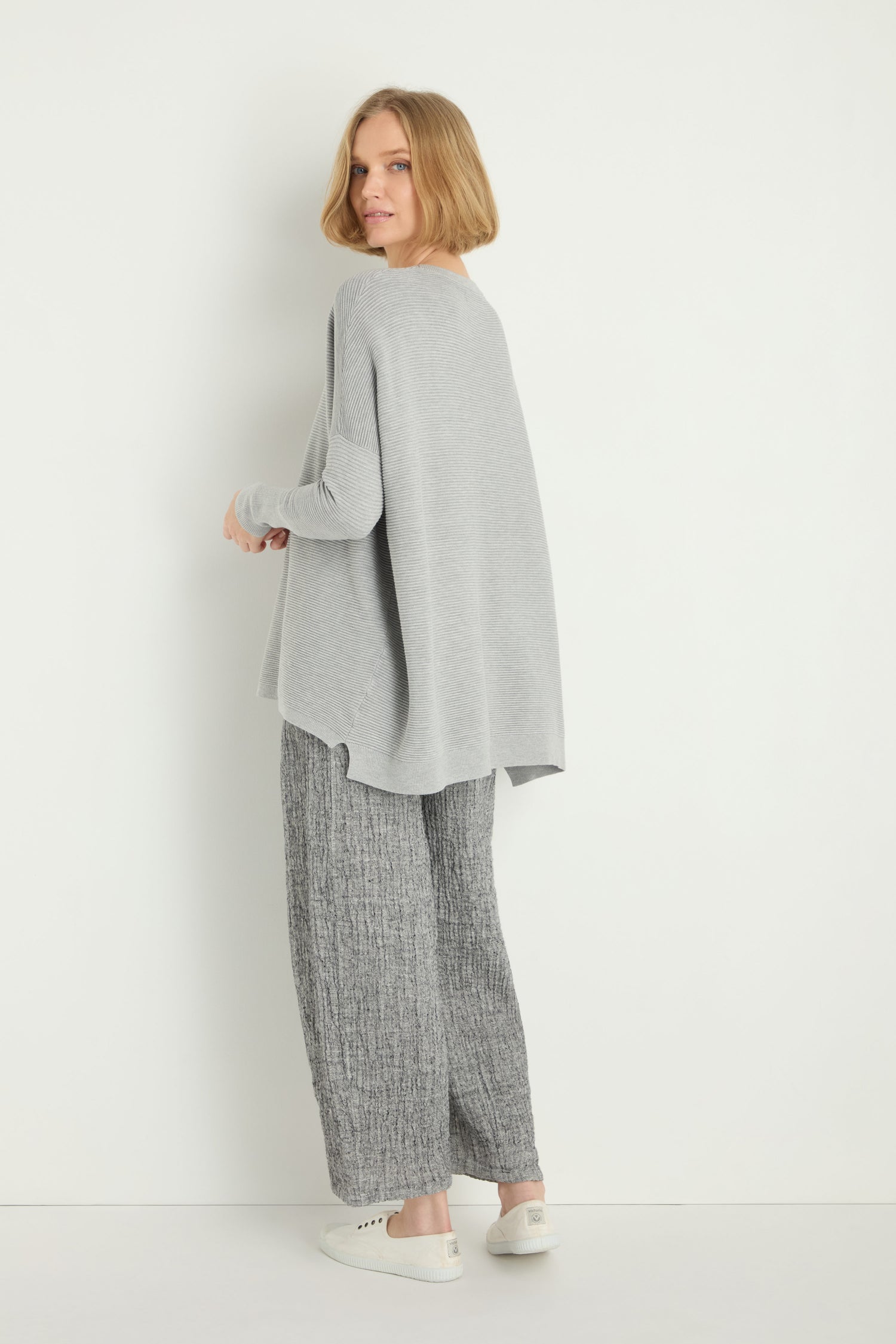 Woman standing against a white wall, wearing a Round Neck Ribbed Top, grey textured pants, and white sneakers, looking over her shoulder at the camera.