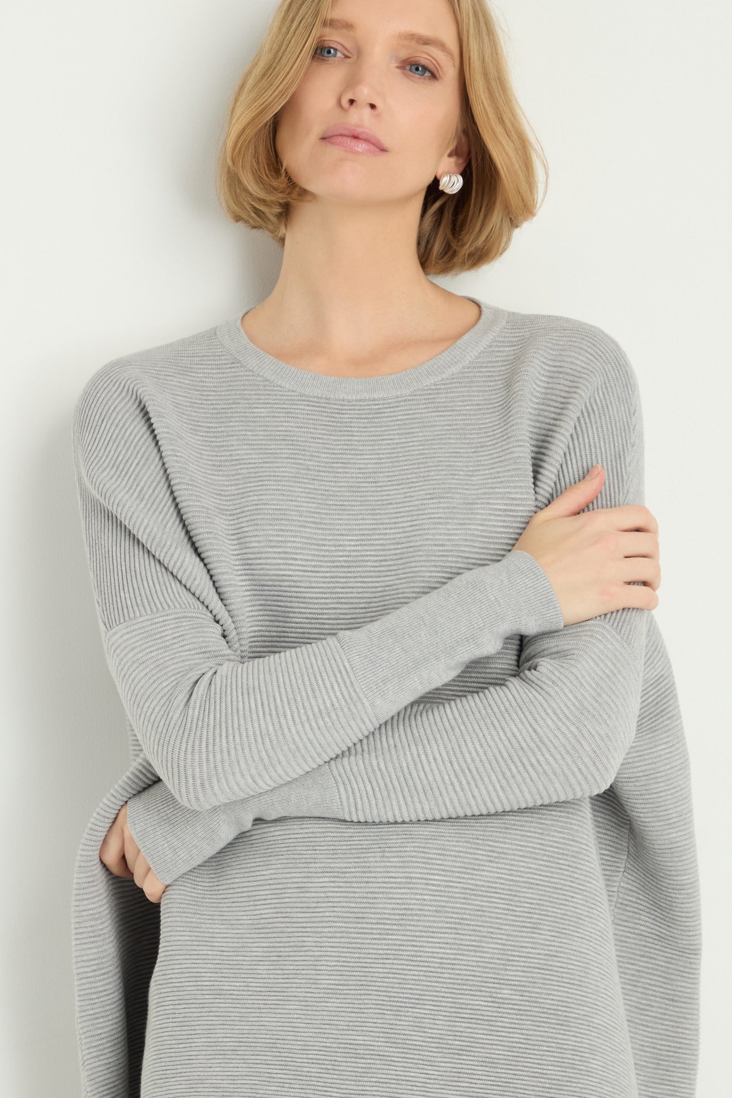A woman with short blonde hair, arms crossed, wears a Round Neck Ribbed Top in light gray, standing against a plain light background.