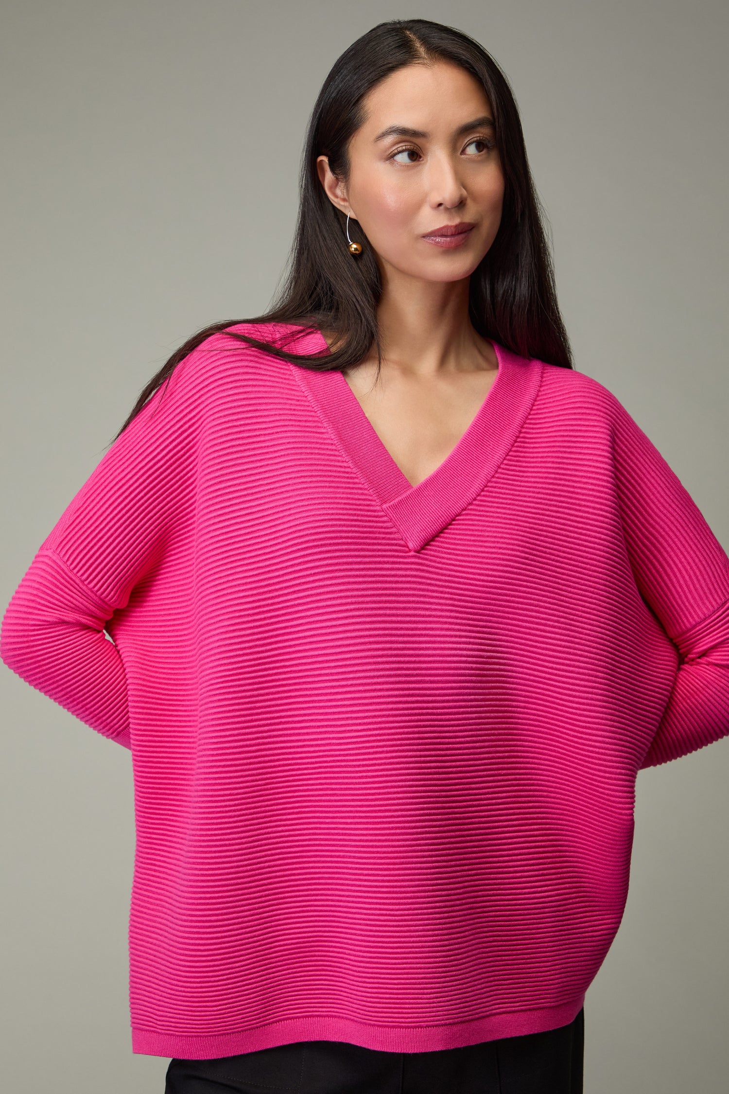 A woman with long dark hair wears the Paisie V Neck Ribbed Jumper in bright pink and black pants, standing against a plain gray background.