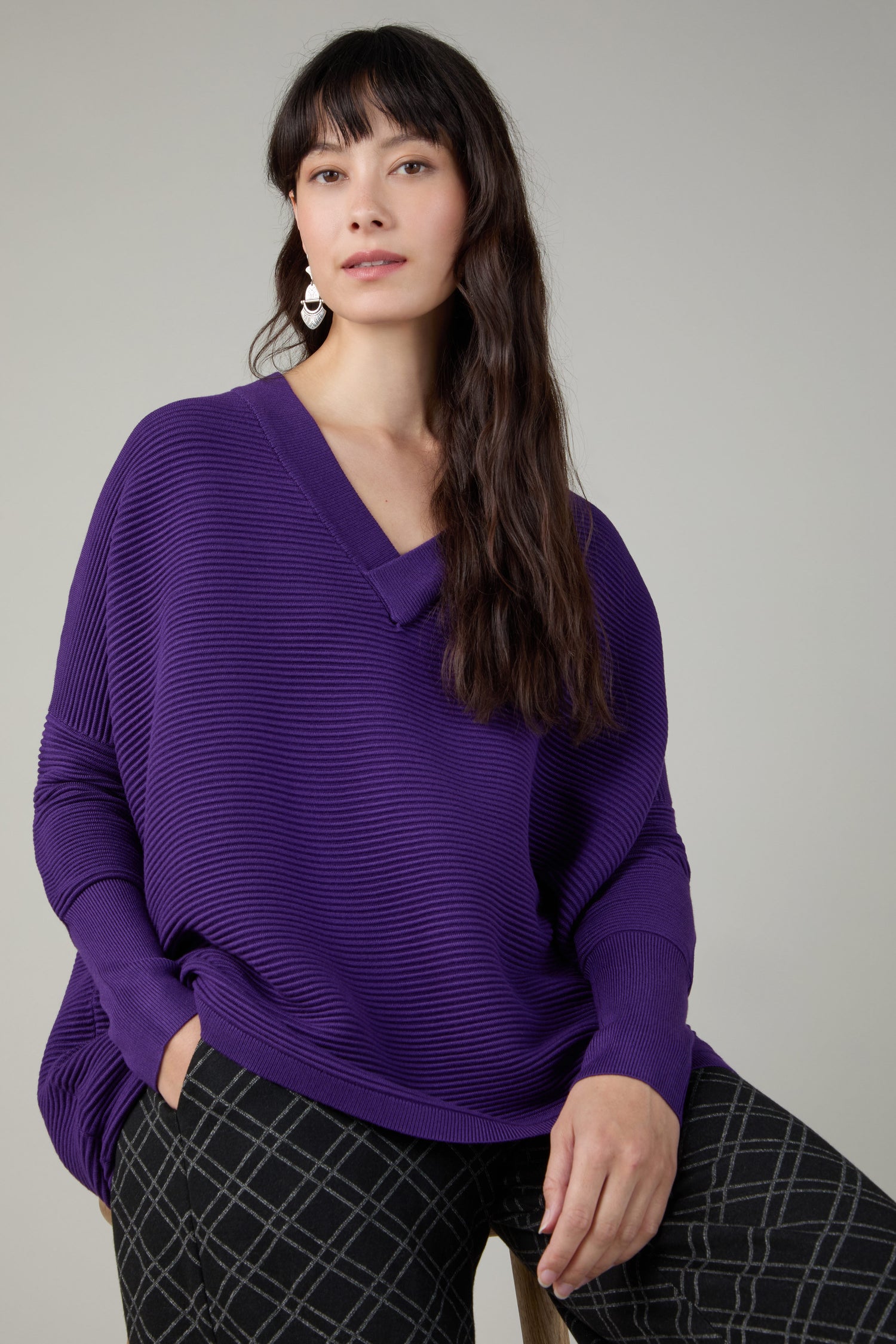 A woman sits on a stool against a plain light gray background, wearing a loose Paisie V Neck Ribbed Jumper in purple and black checkered pants.