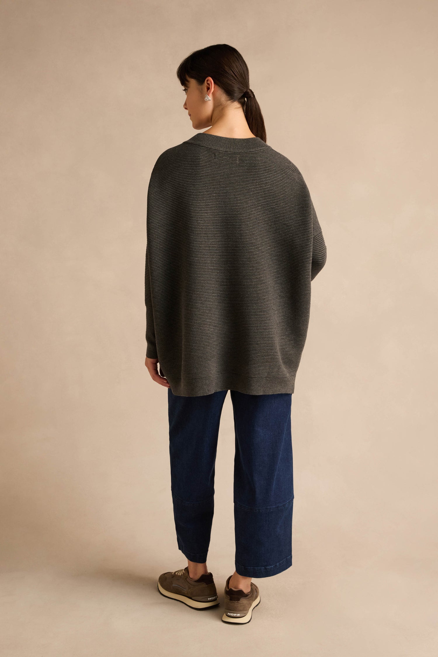 A person stands facing away, wearing the Charcoal V Neck Ribbed Jumper—a relaxed fit piece crafted from a soft viscose blend—paired with wide-leg blue jeans and brown sneakers, hair pulled back in a ponytail, against a plain beige background.