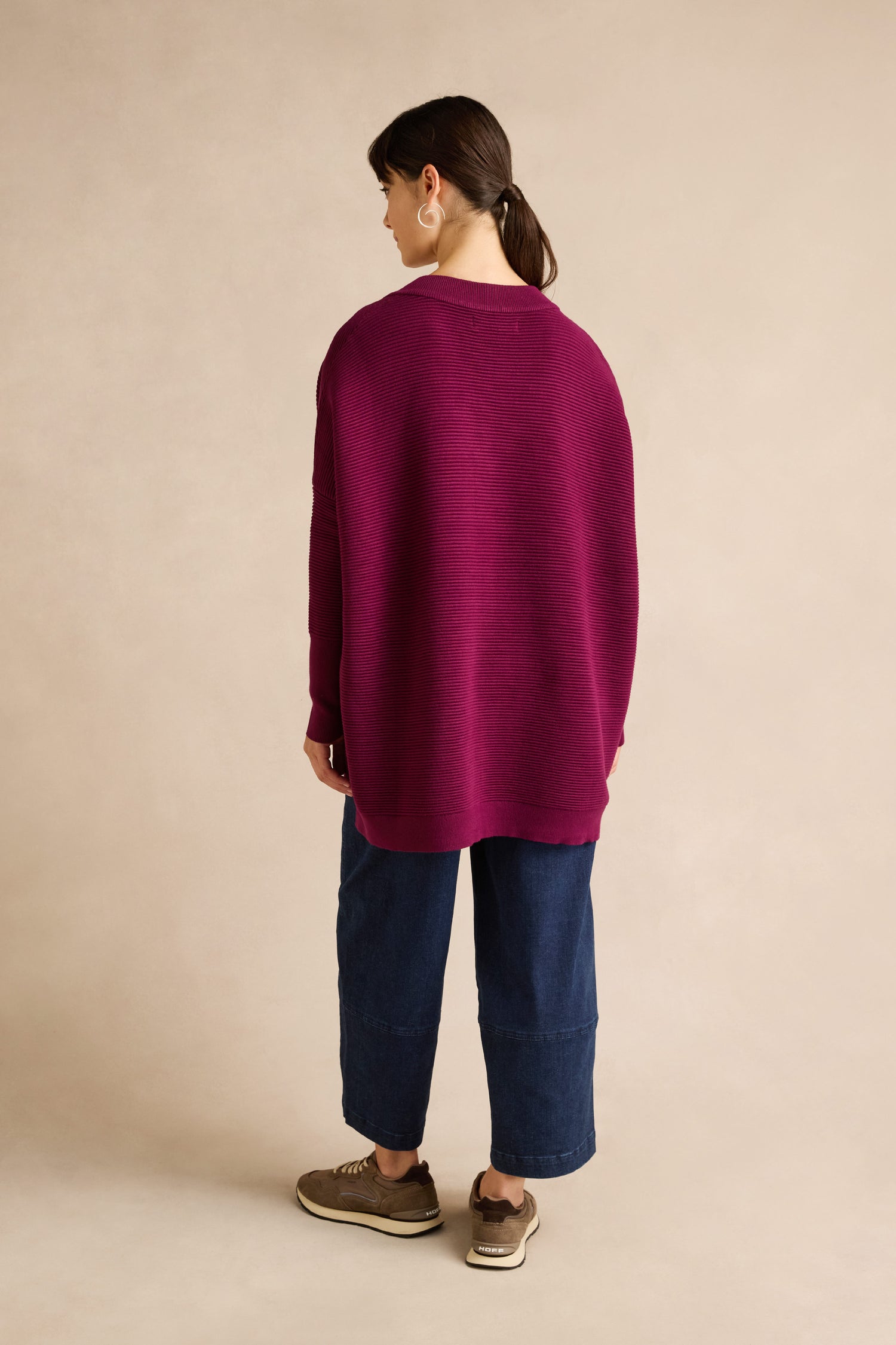 A person stands facing away, wearing the Wine V Neck Ribbed Jumper in magenta crafted from a soft viscose blend with a relaxed fit and ribbed design, paired with wide-leg blue jeans and brown shoes against a plain beige background.