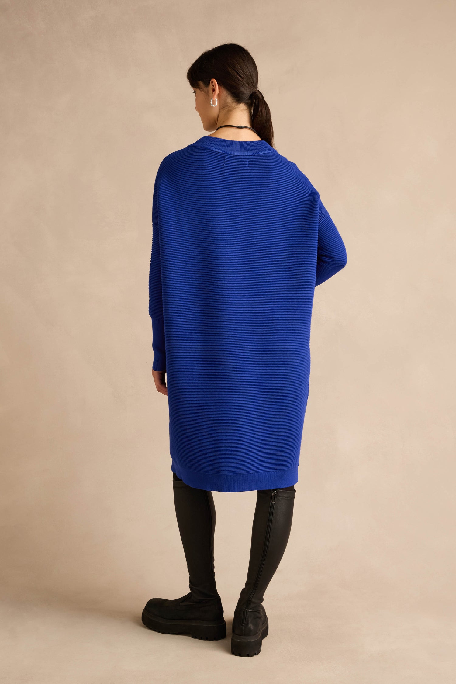 A person stands with their back to the camera, wearing the V Neck Ribbed Dress in royal blue, styled with black leggings and black shoes against a neutral background.
