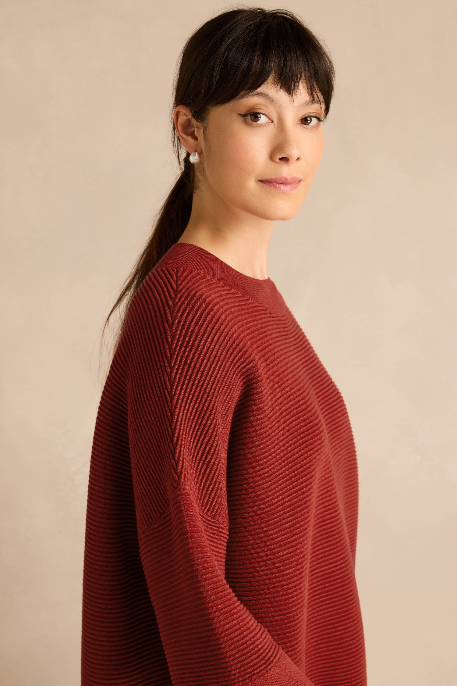 A woman with straight dark hair in a ponytail, wearing the Wide Sleeve Burgundy Ribbed Knit sweater, stands against a plain beige background, gazing slightly to the side.
