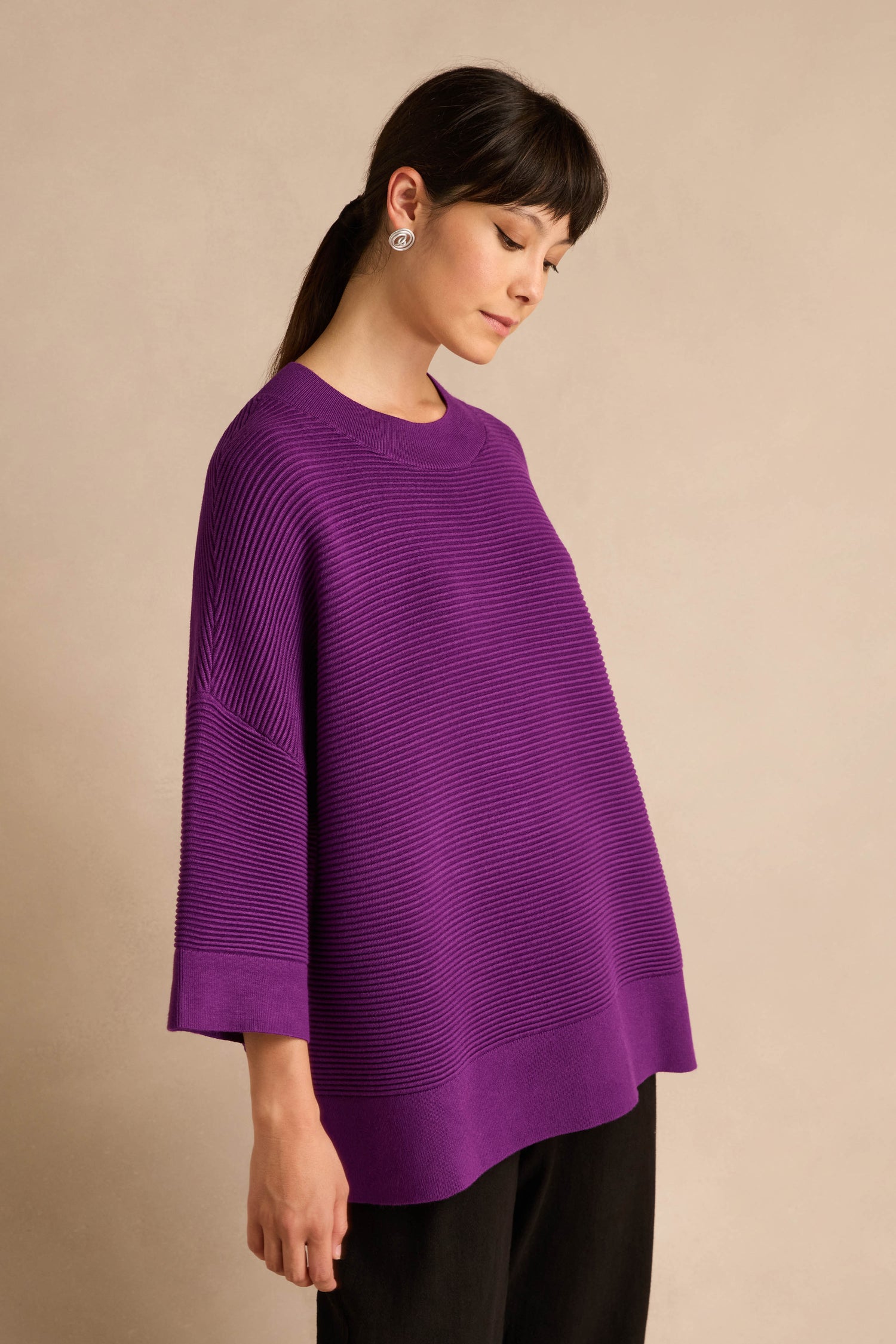 A woman stands against a beige background wearing the Wide Sleeve Purple Ribbed Knit sweater paired with black pants; her hair is tied back and she finishes the look with silver earrings.