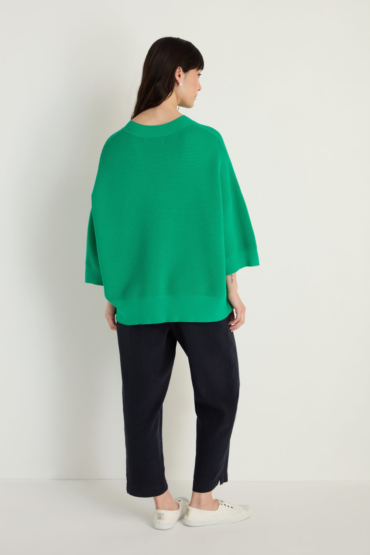 A woman faces away, wearing the Wide Sleeve Ribbed Top in green with ribbed texture, paired with black pants and white sneakers, set against a plain light background.