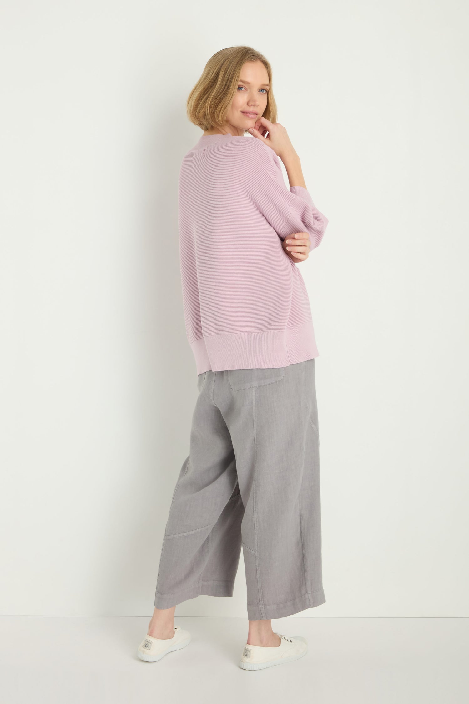 A woman stands against a white wall wearing the Wide Sleeve Ribbed Top in light purple, paired with gray wide-leg pants and white slip-on shoes, glancing over her shoulder.