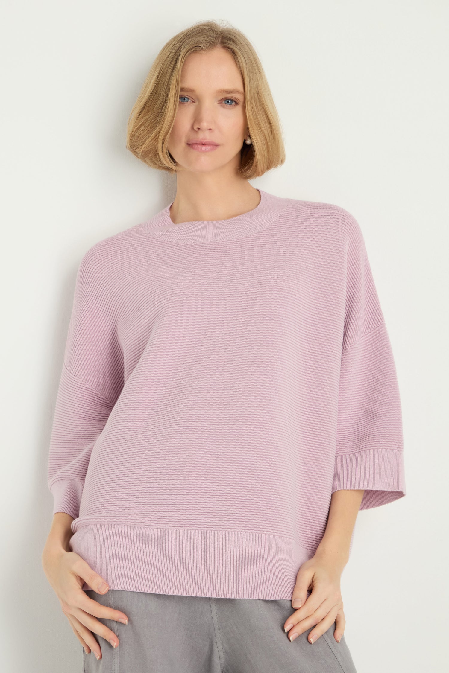A woman with short blonde hair wears the Wide Sleeve Ribbed Top in light lavender with a ribbed texture and gray pants, standing against a plain white background.